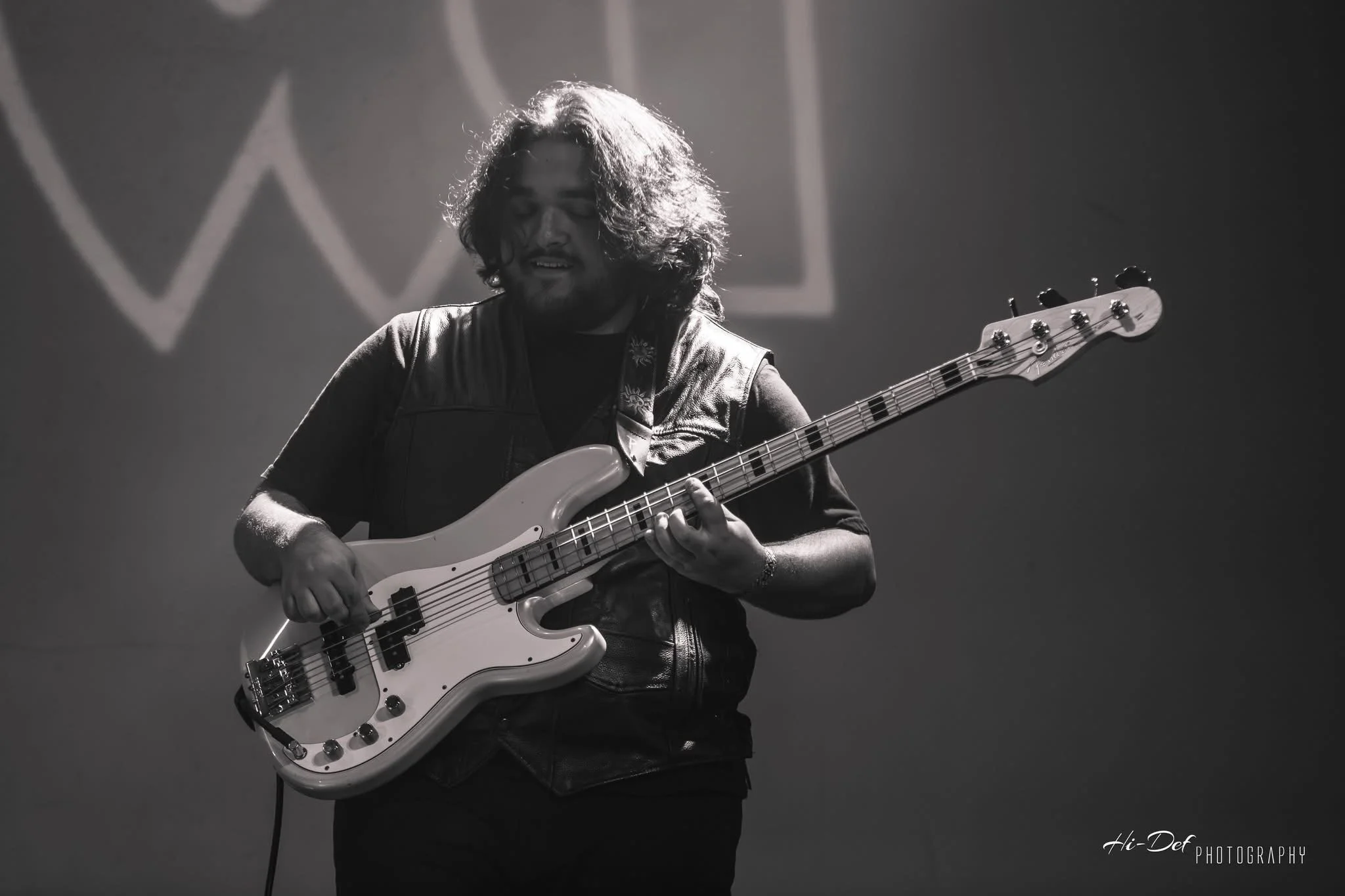 A man playing a bass guitar during a live performance, illuminated by stage lighting, with a dark background and a logo in the background.