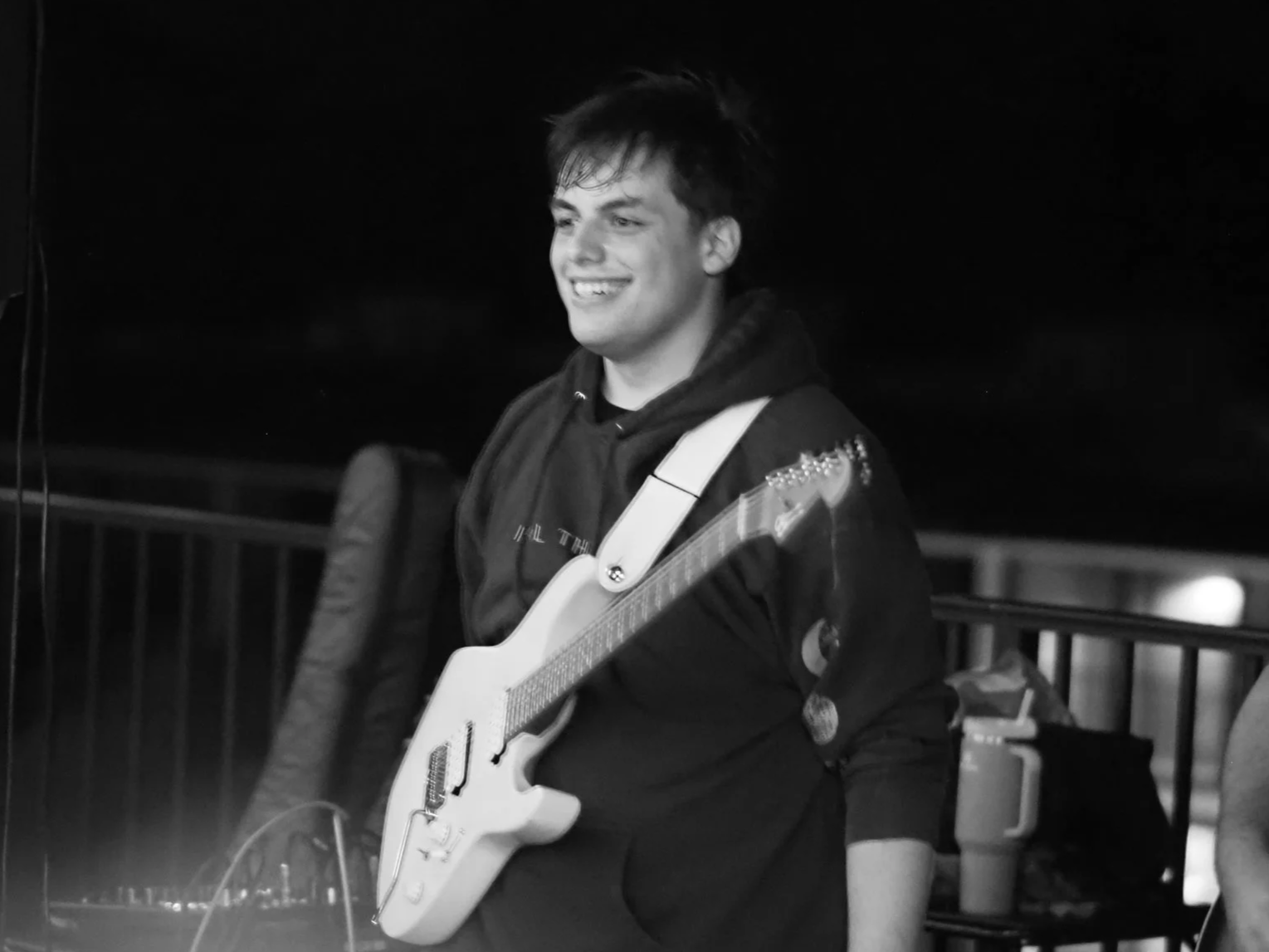 A young man smiling while holding an electric guitar, wearing a hoodie and standing near music equipment at night.