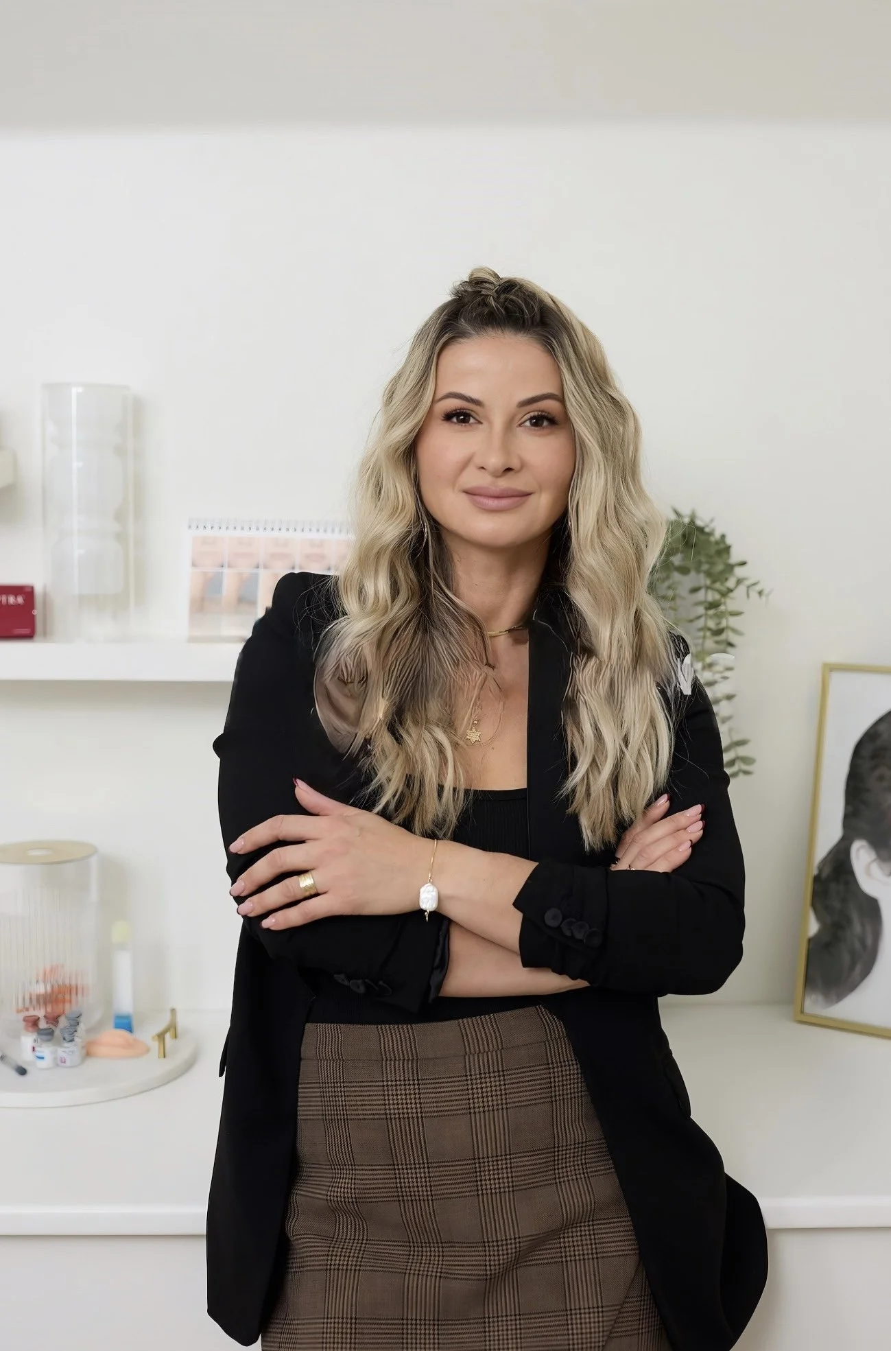 A woman with long wavy blonde hair, wearing a black blazer and a brown plaid skirt, standing with her arms crossed in an office or clinic setting.