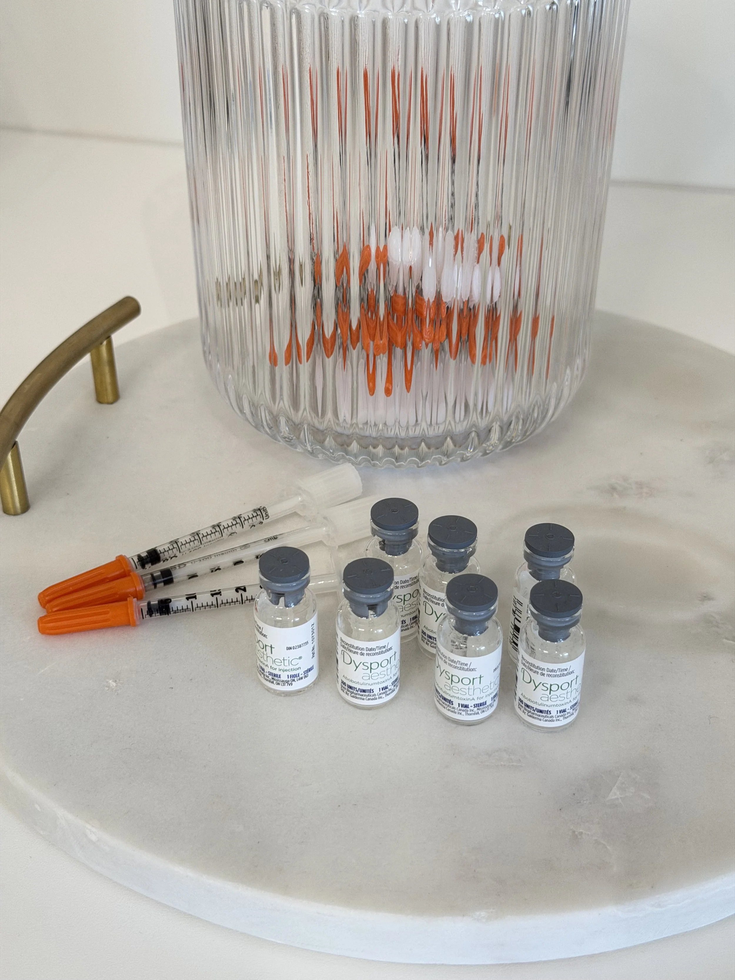 Medical syringes and vials labeled Dysport on a white surface with a large glass container in the background.