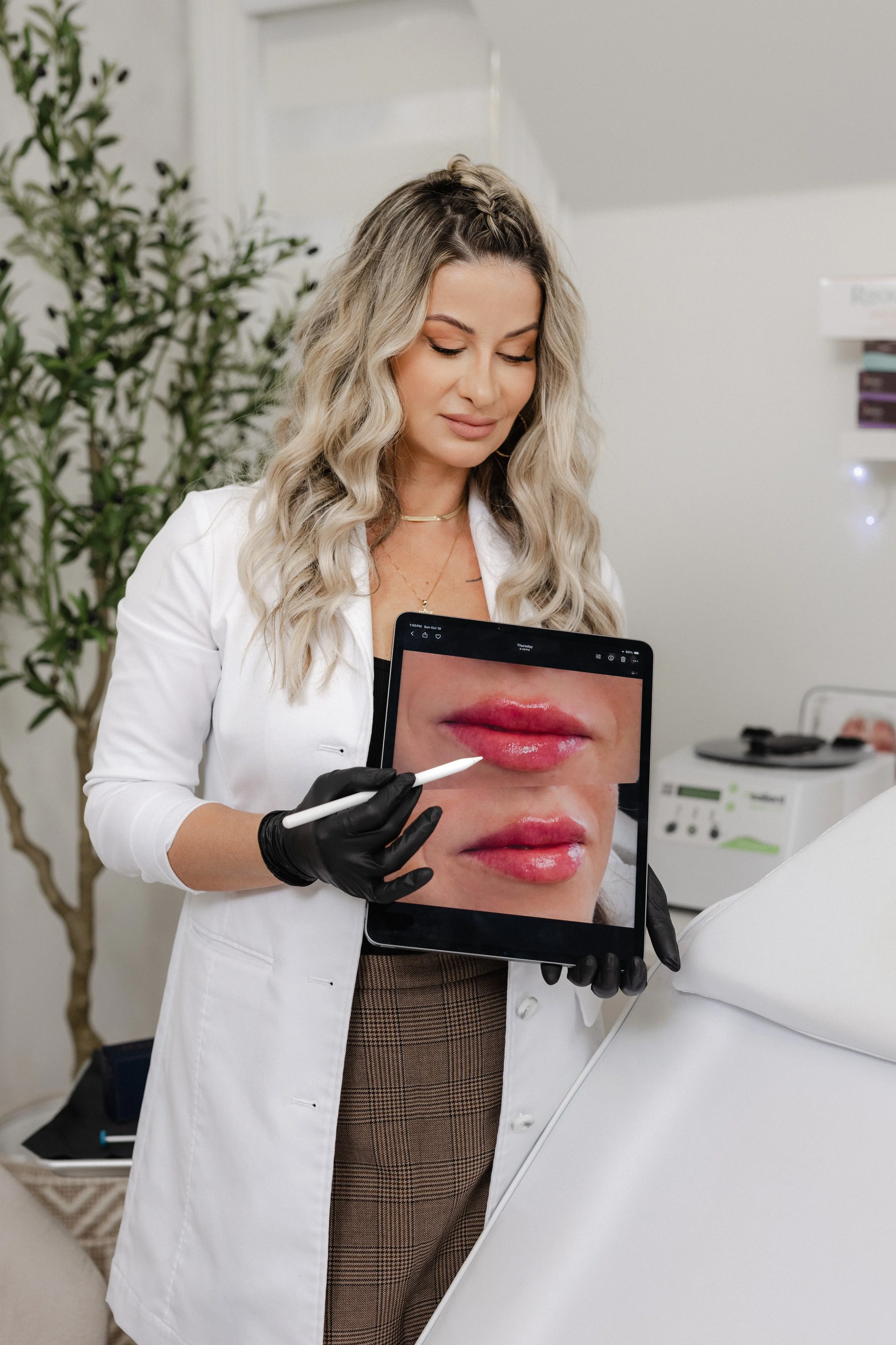 A woman in a white coat and black gloves is holding a tablet showing a close-up image of lips, at a medical or beauty clinic.