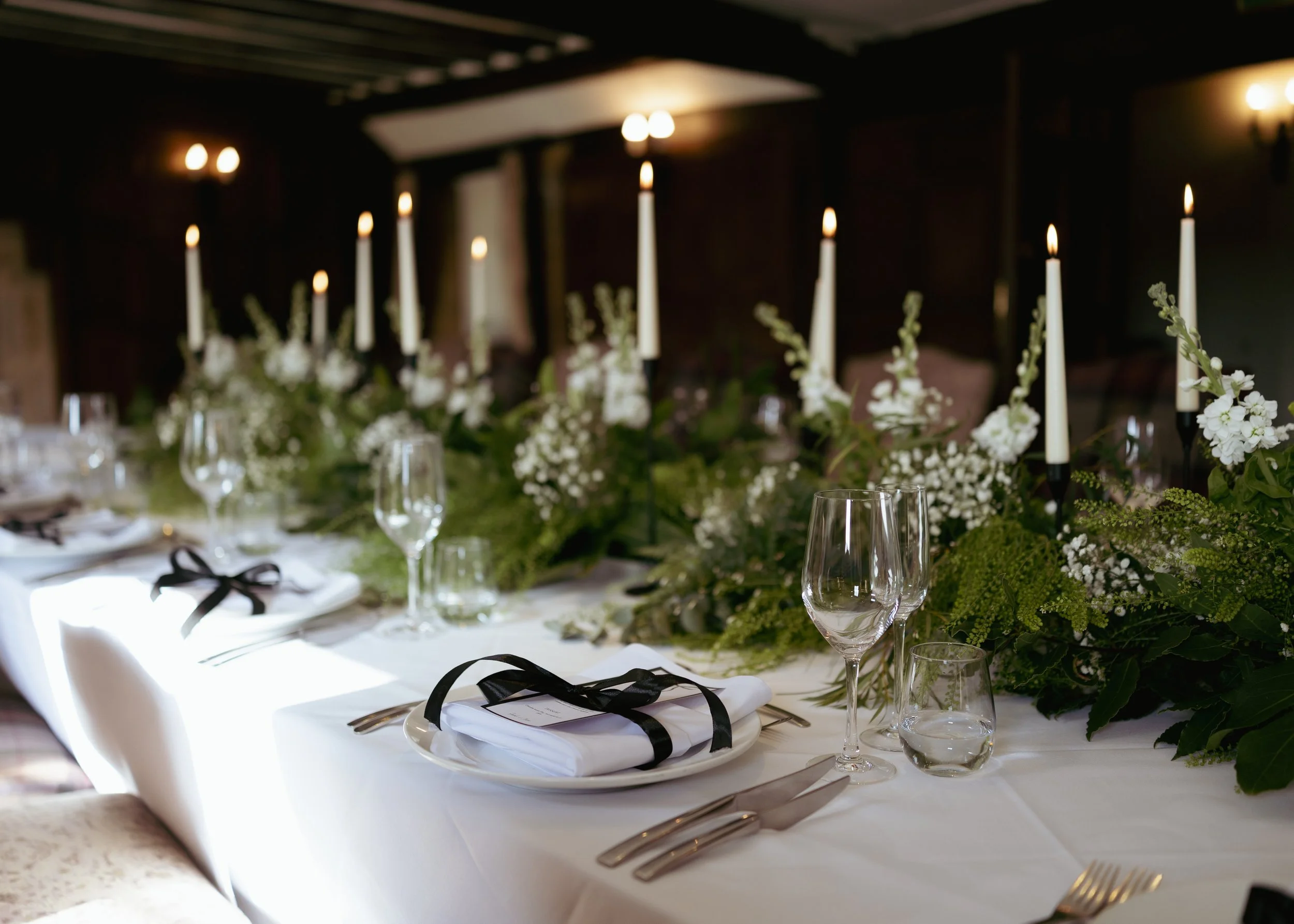Elegant dining table setup with white tablecloth, wine glasses, silverware, and black ribbon-tied napkins. The table features tall white candles and floral arrangements with white flowers and greenery in a dimly lit room.