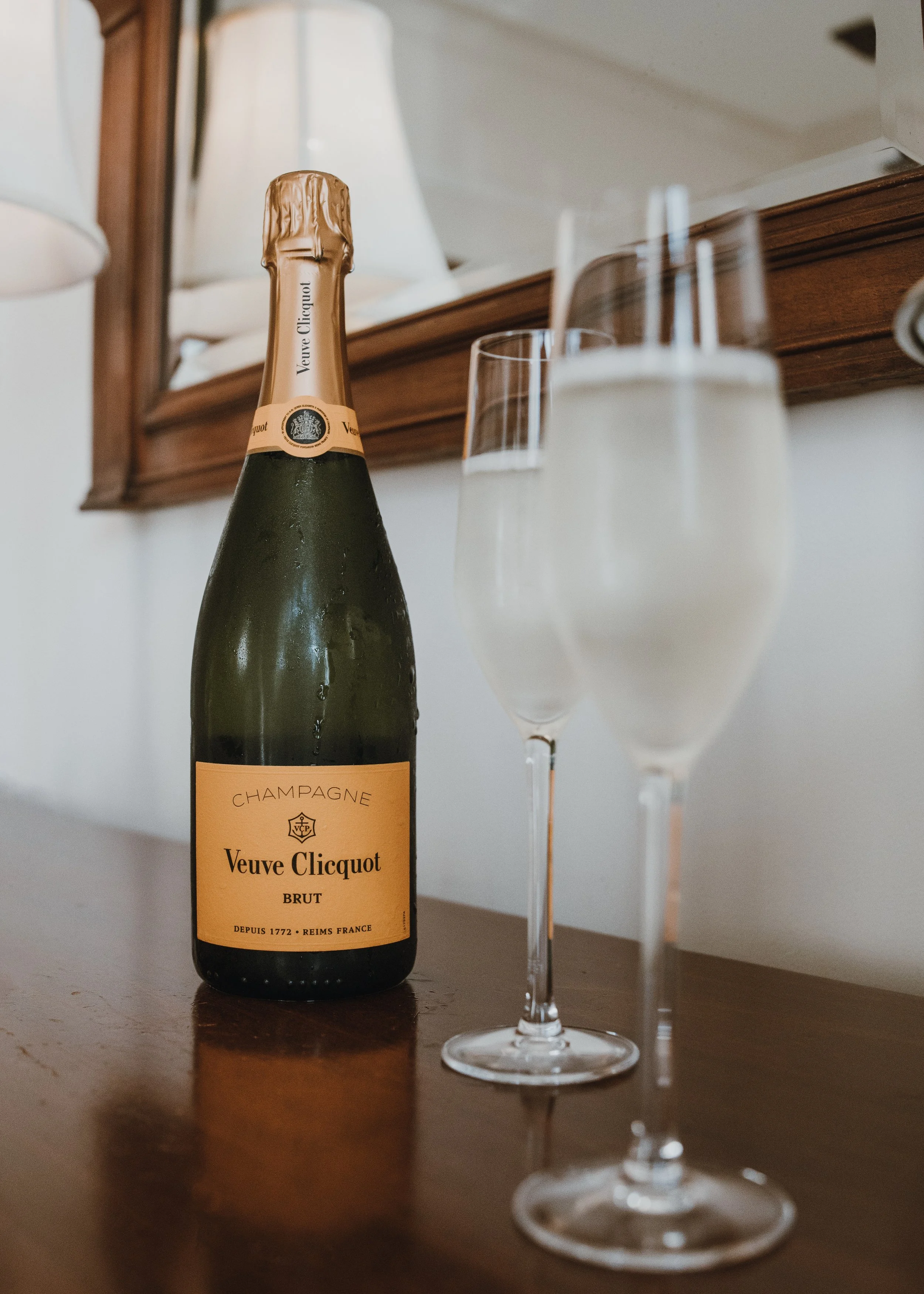 A bottle of Veuve Clicquot champagne and two filled champagne flutes on a wooden surface against a white wall with a mirror and lamps in the background.