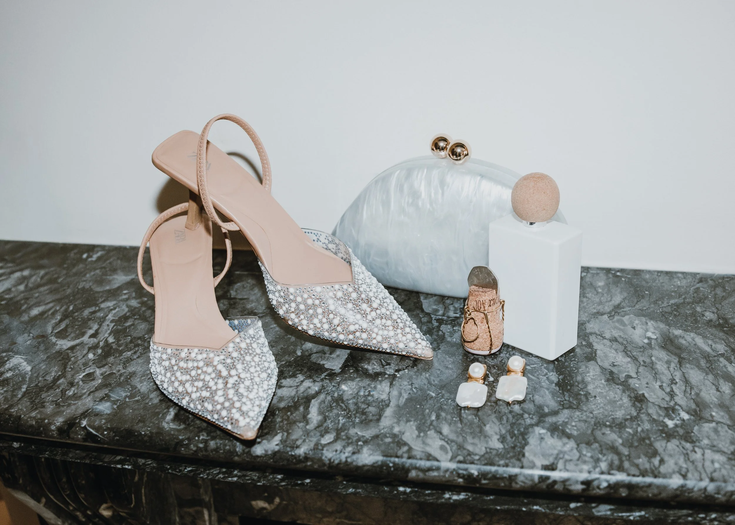 Beige high-heeled shoes adorned with pearls and beads, jewelry including earrings and rings, and decorative objects on a dark marble surface.