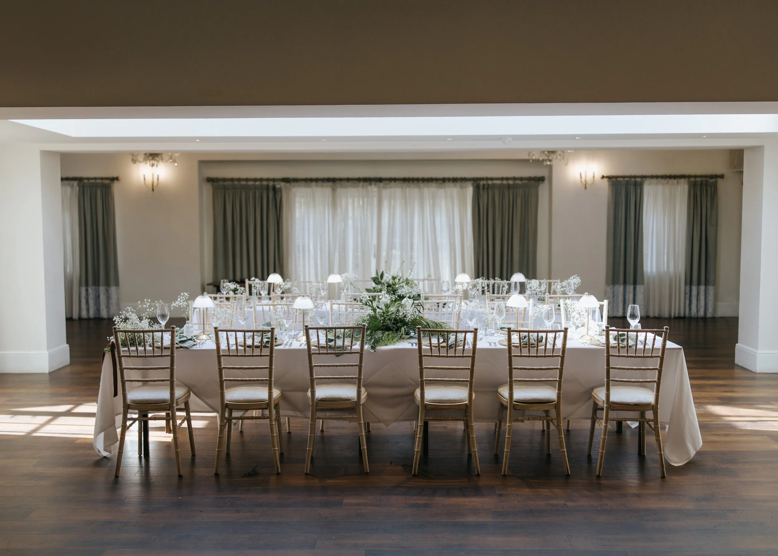 Elegant banquet table set up in a bright dining room with floral centerpiece, white tablecloth, and arranged glassware and plates, surrounded by wooden chairs. Large windows with curtains allow natural light to fill the room.