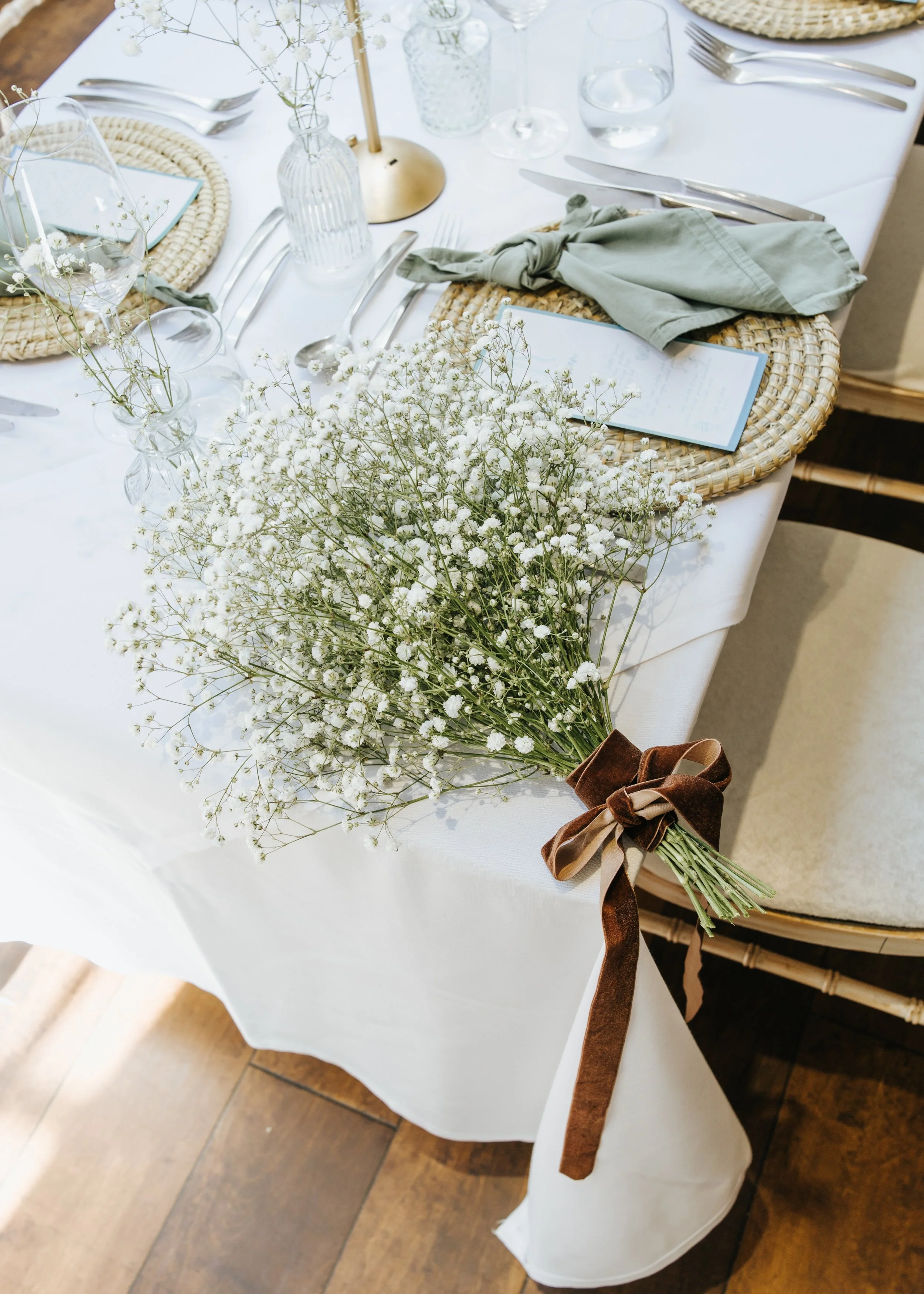 Table decorated with a large bouquet of white baby's breath flowers tied with a brown ribbon, set with woven placemats, menus, silverware, water glasses, and small vases with more baby’s breath. The table is covered with a white tablecloth.