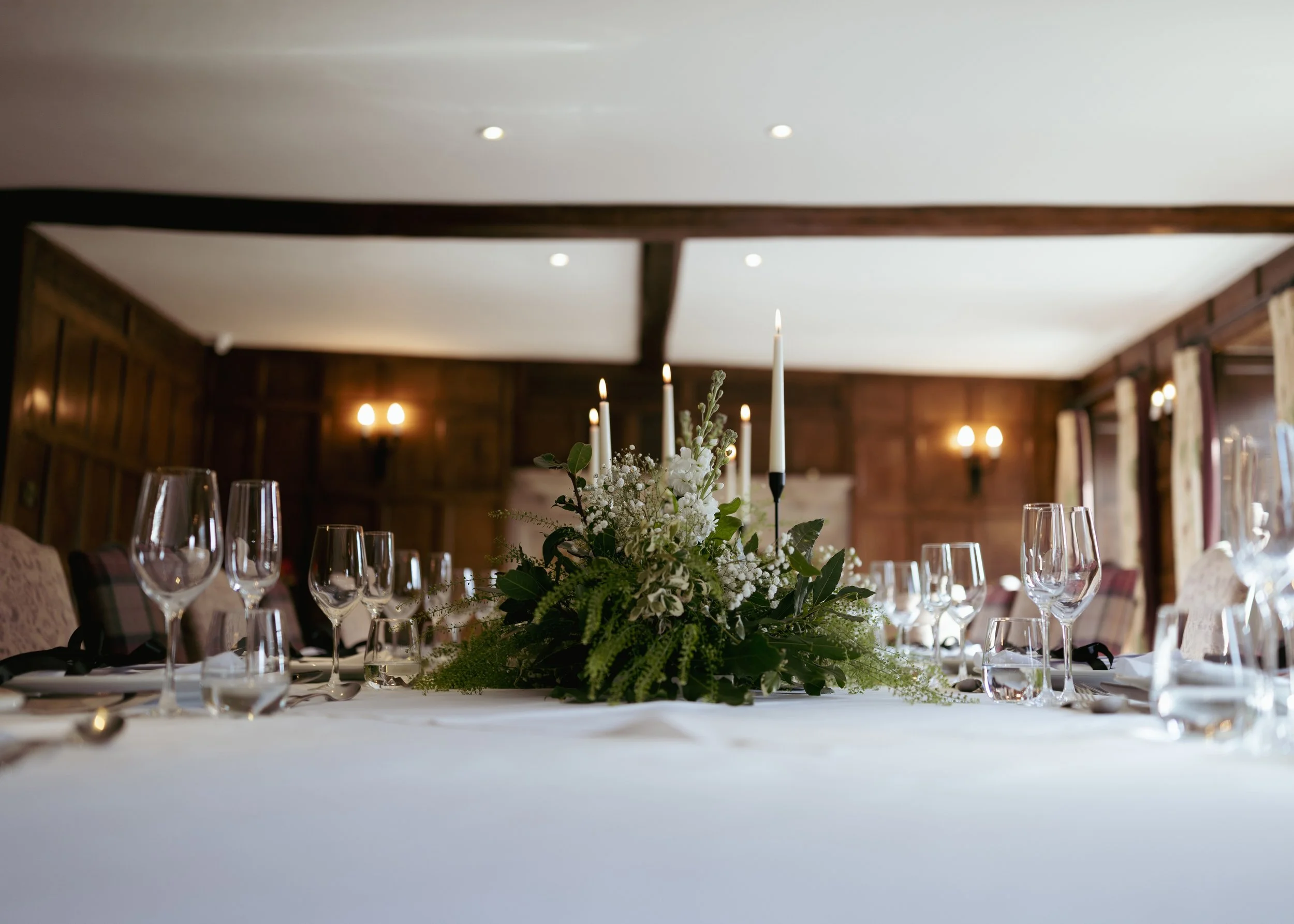 Elegant banquet table set with wine glasses, silverware, and a floral centerpiece with candles in a wood-paneled room.