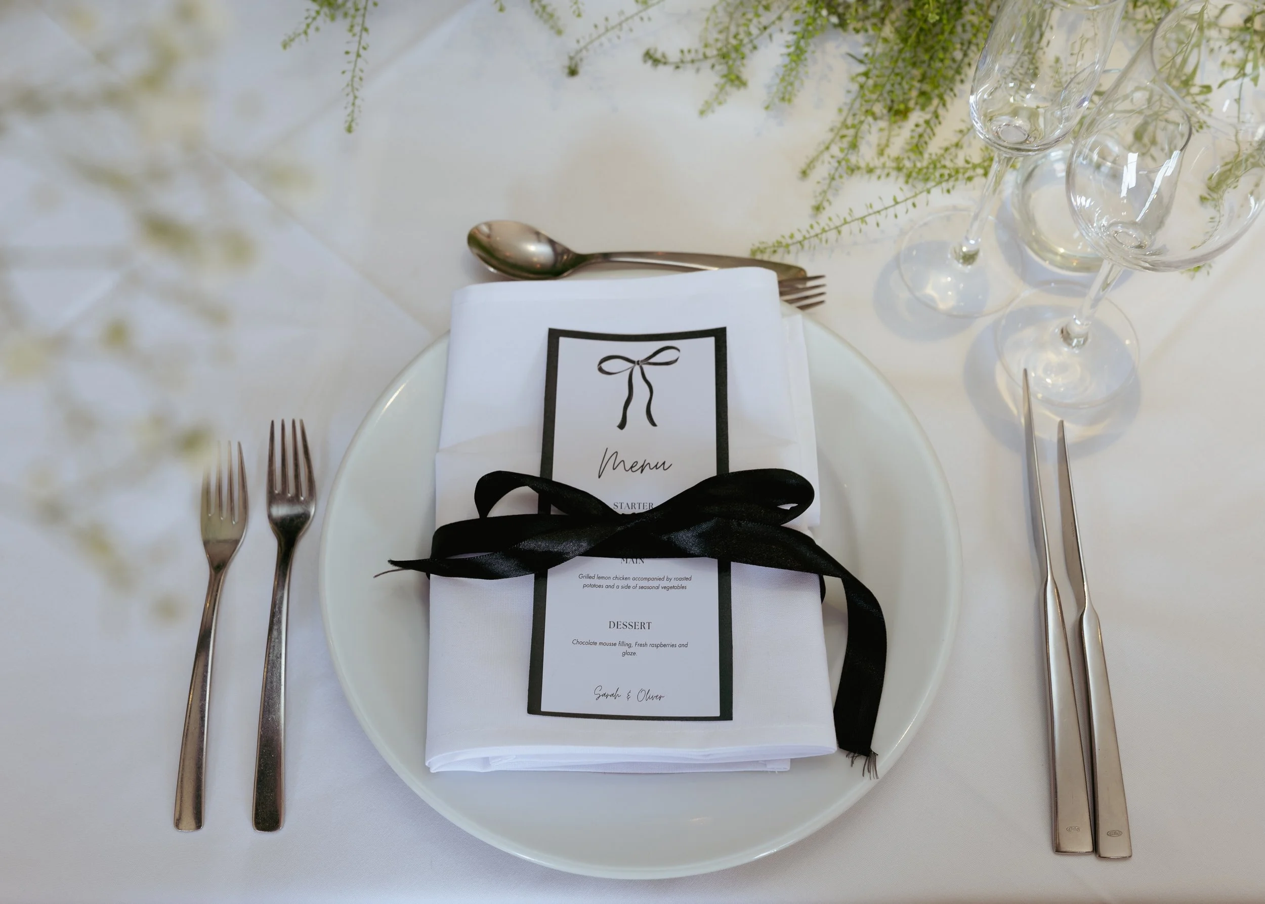 Elegant table setting with a white plate, silverware, wine glasses, and a neatly folded napkin tied with a black ribbon. A printed menu is placed on top of the napkin.