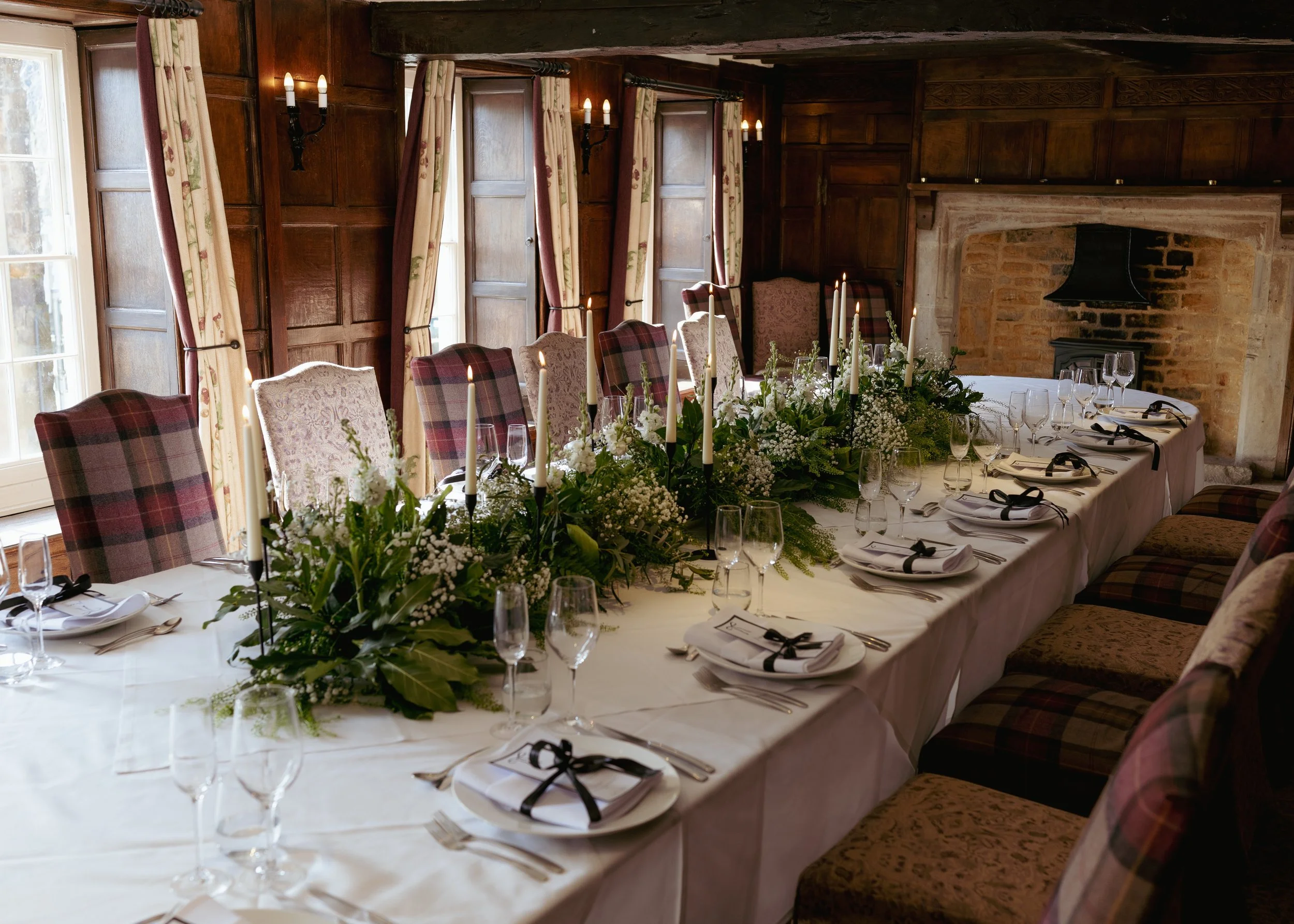 A long dining table set for a formal event with white tablecloth, floral centerpiece, candles, and arranged with plates, silverware, wine glasses, and napkins tied with black ribbons. The room features wood-paneled walls, windows with floral curtains
