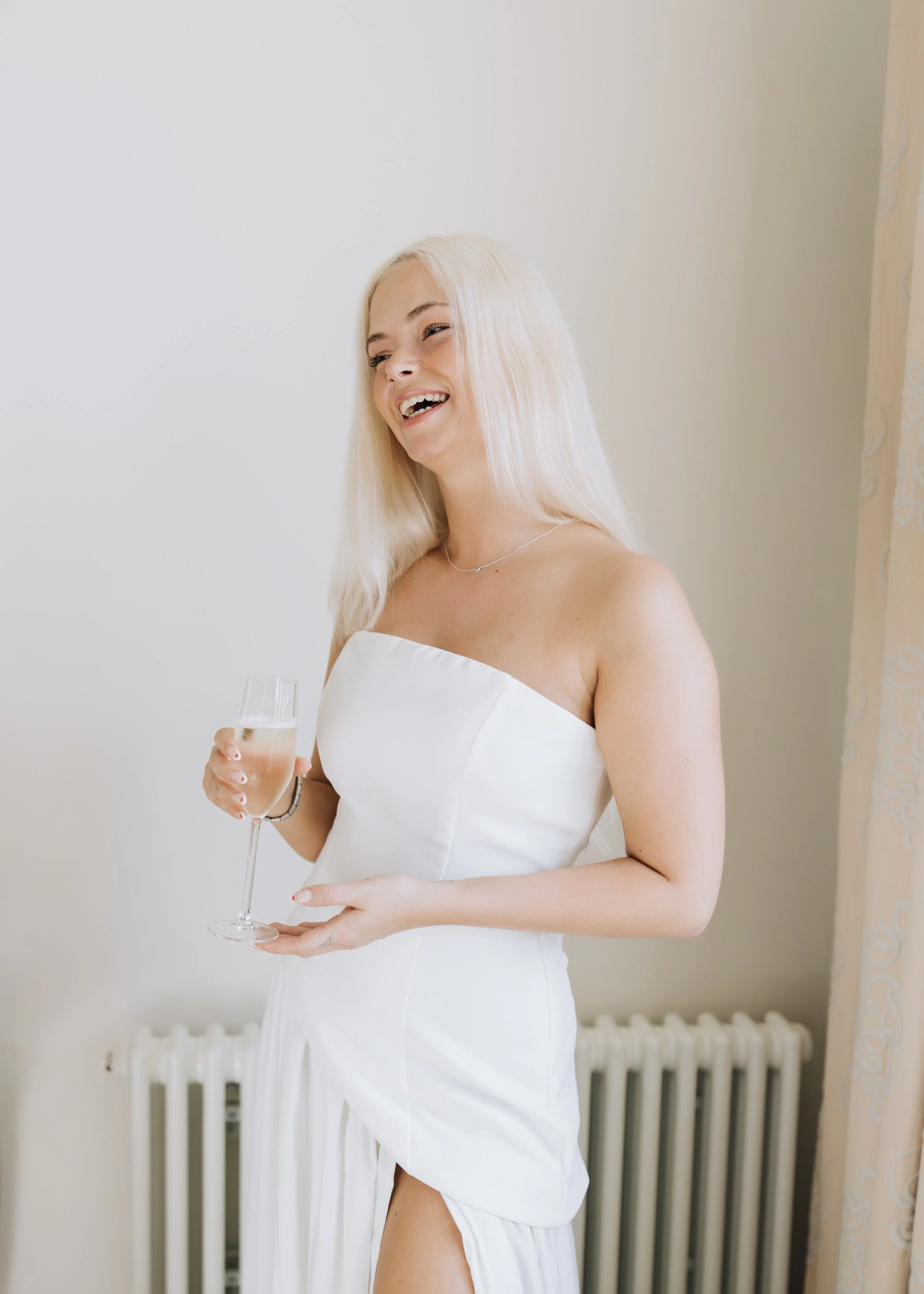 A woman with long blonde hair, wearing a strapless white dress, holding a glass of champagne, and smiling.