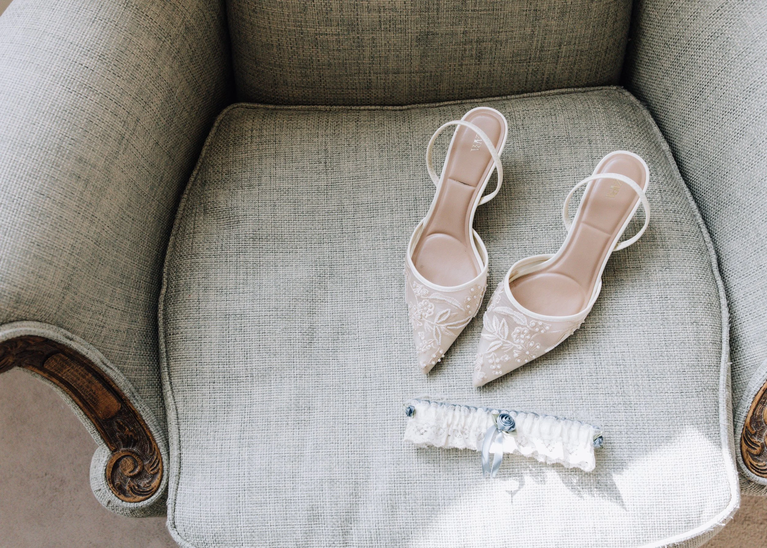 A pair of white satin bridal shoes with embroidery and a matching garter on a vintage sofa.