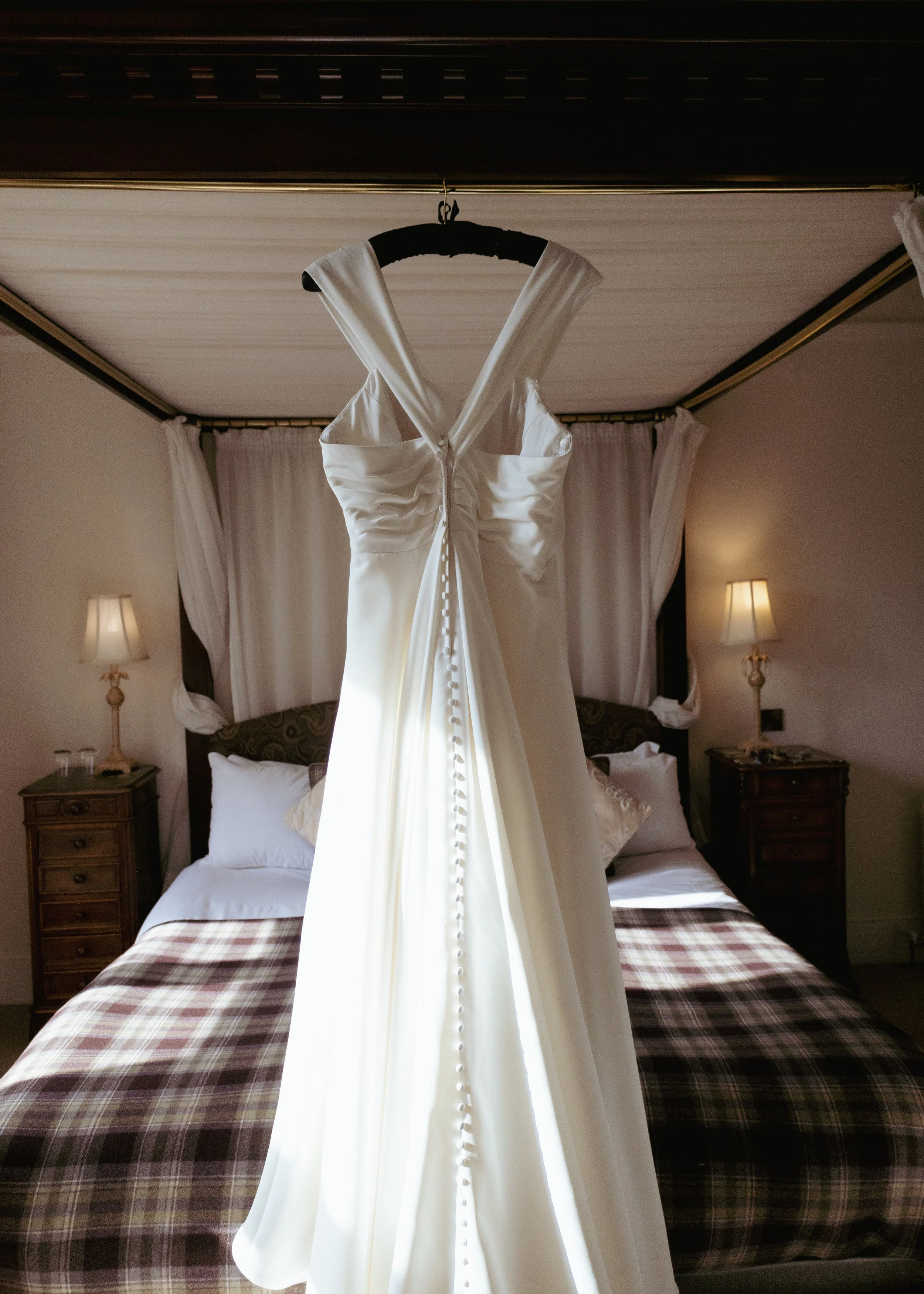 A white wedding dress hanging on a black hanger in a bedroom with two wooden nightstands and wall-mounted lamps.