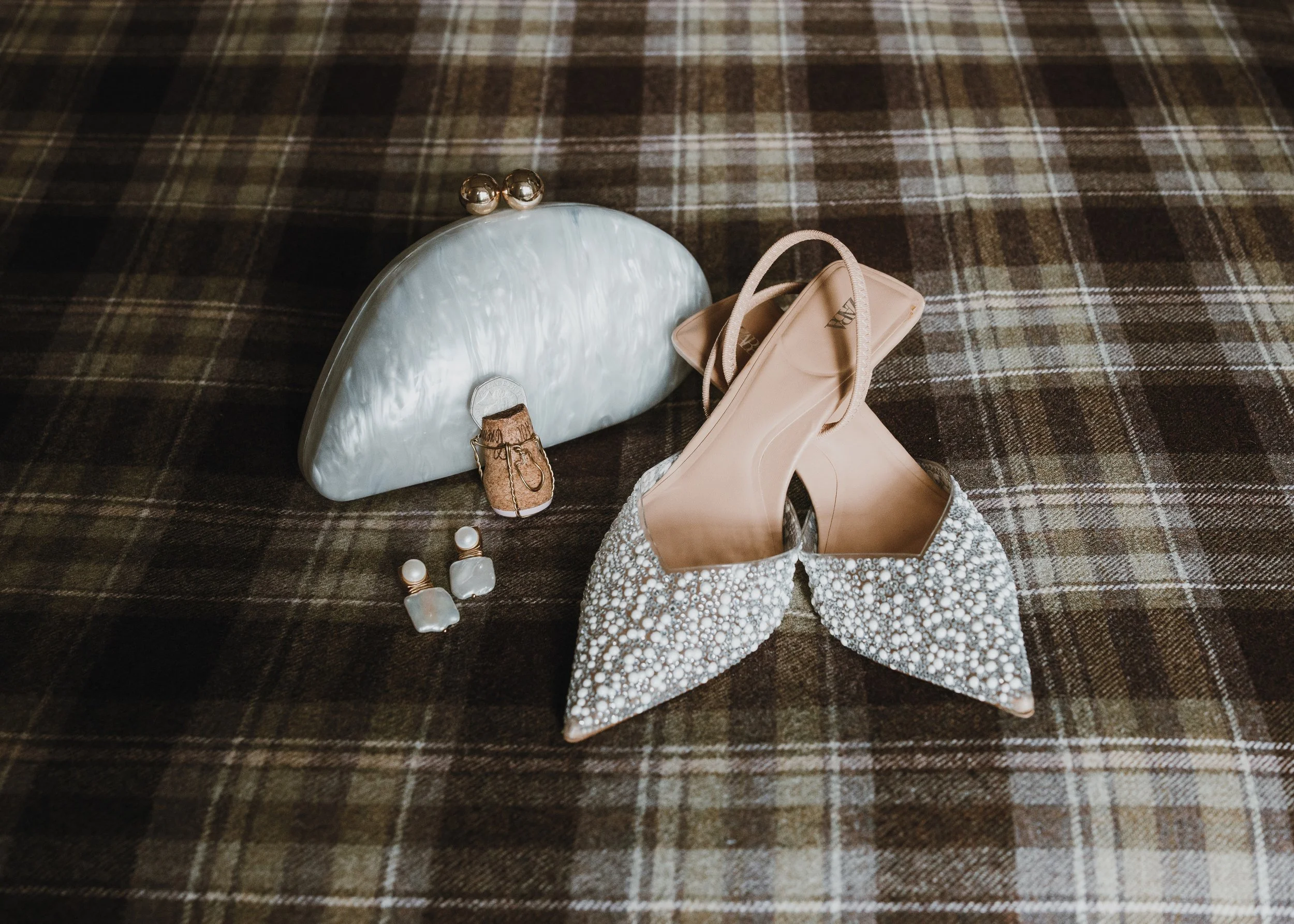 A collection of elegant fashion accessories on a brown plaid fabric surface, including a pearl and mother-of-pearl clutch, a pair of white beaded high-heeled shoes, pearl and clear gemstone earrings, a small pearl and gold ring, and a miniature shoe 