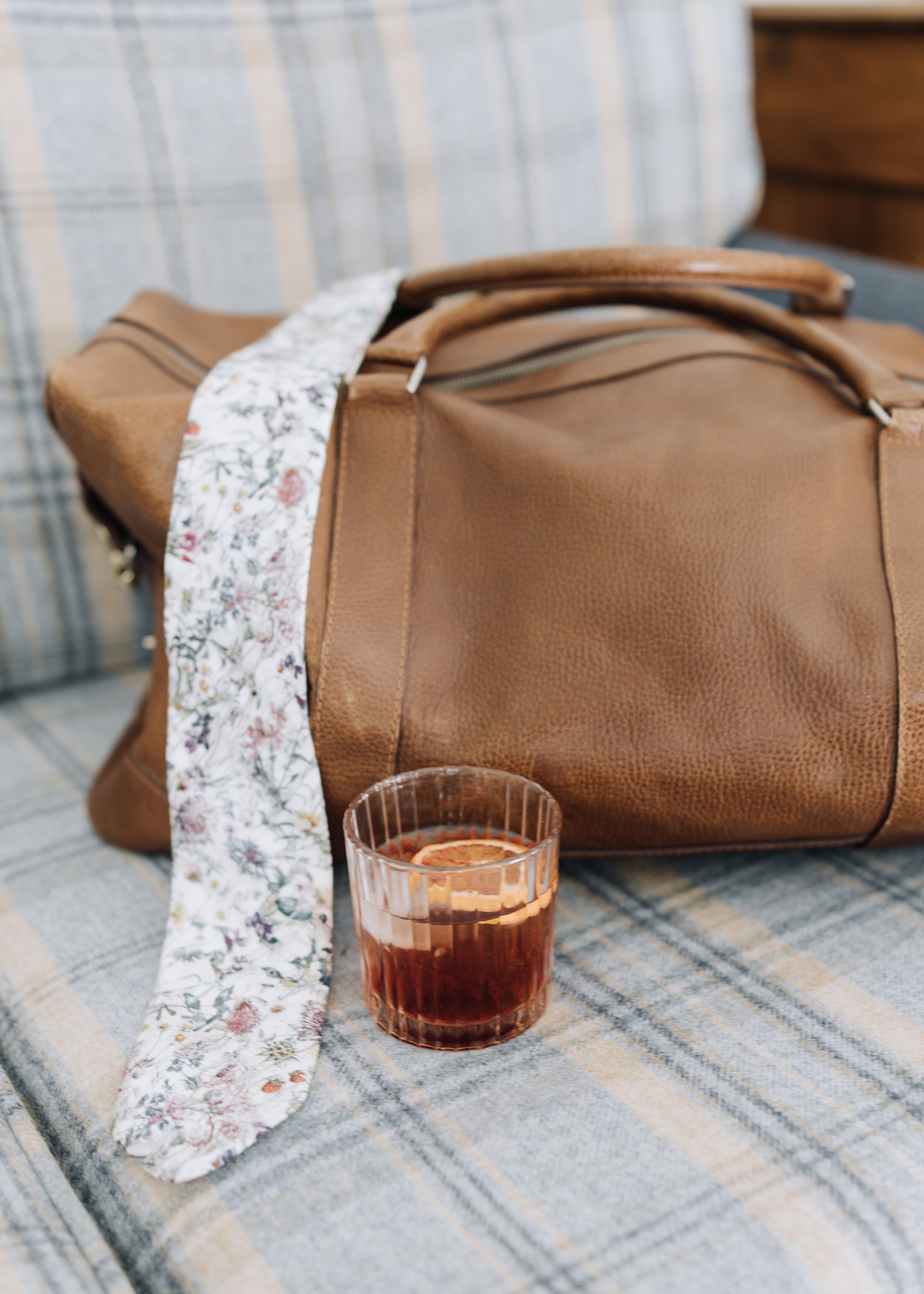 A brown leather duffel bag with a floral-patterned strap on a plaid upholstered bench, next to a glass of dark-colored beverage with a lime slice.