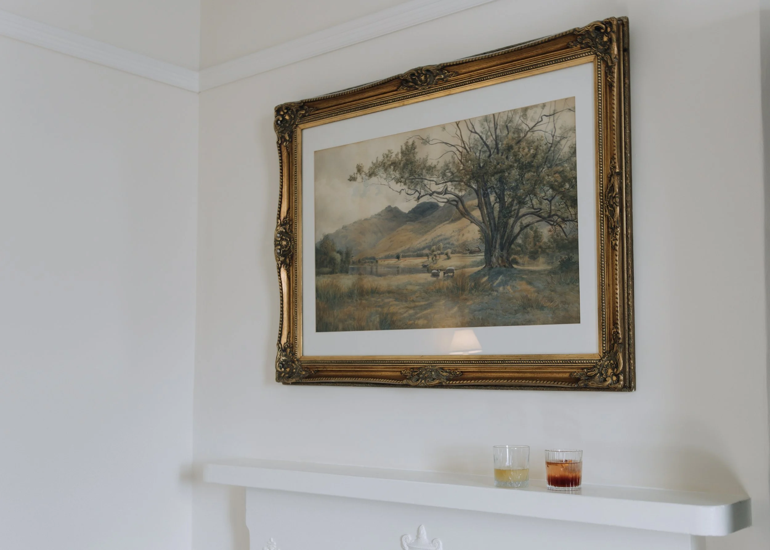 A framed landscape painting of a tree, mountains, and a river hangs on a white wall. Below, on a white mantel, are two glasses, one with a yellow drink and the other with a reddish drink.