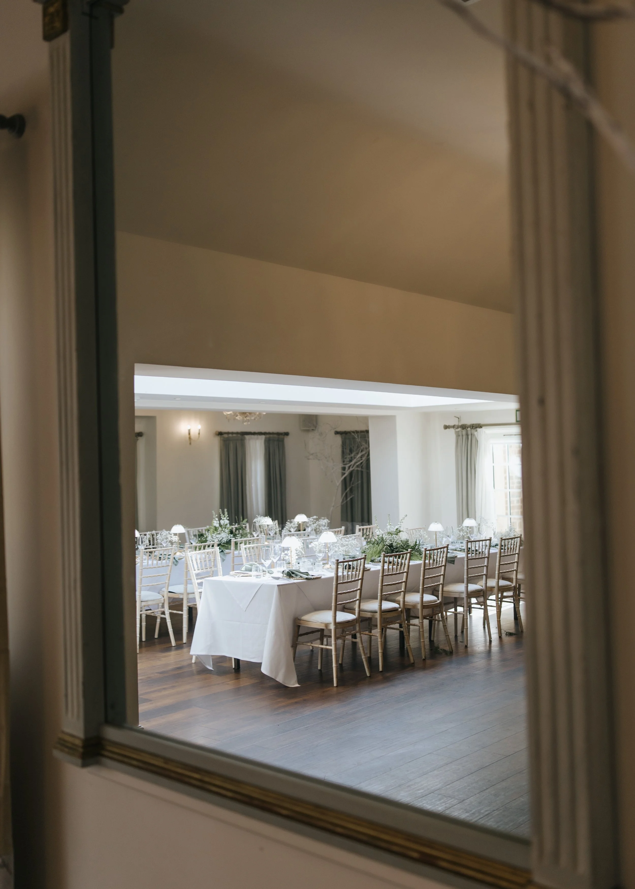 Empty banquet hall viewed through a mirror, decorated for an event with white tablecloths, floral centerpieces, and gold chairs.