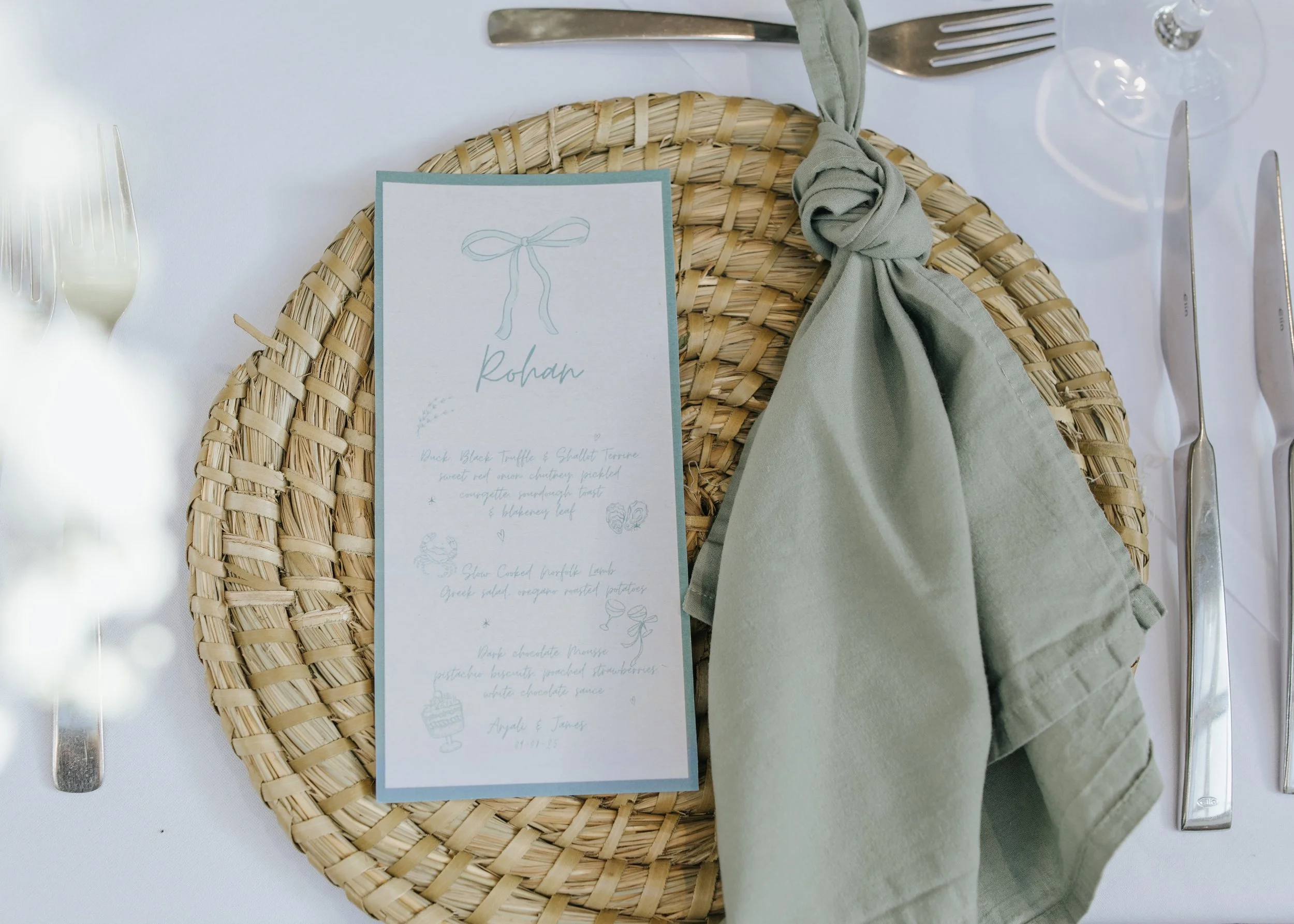 A place setting with a woven round placemat, a green napkin knotted around a corner, and a menu card with light blue accents and handwritten-style text.