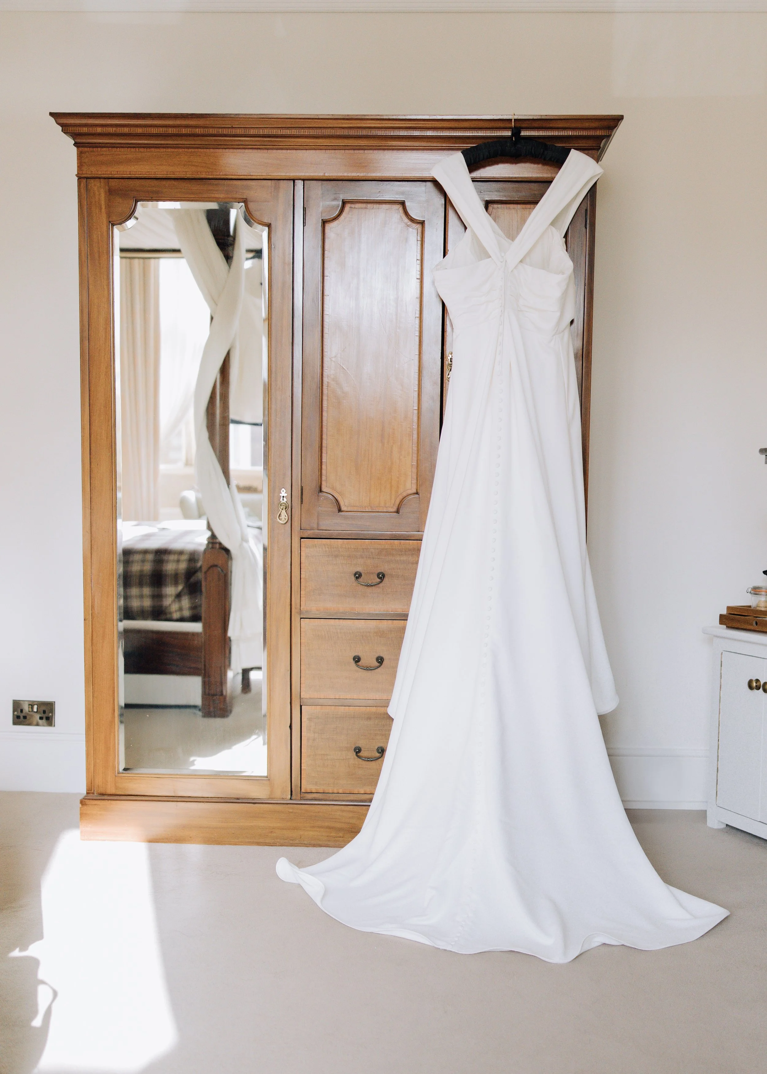 A white wedding dress hanging on a black hanger from the top of a wooden wardrobe with mirror doors in a bedroom.