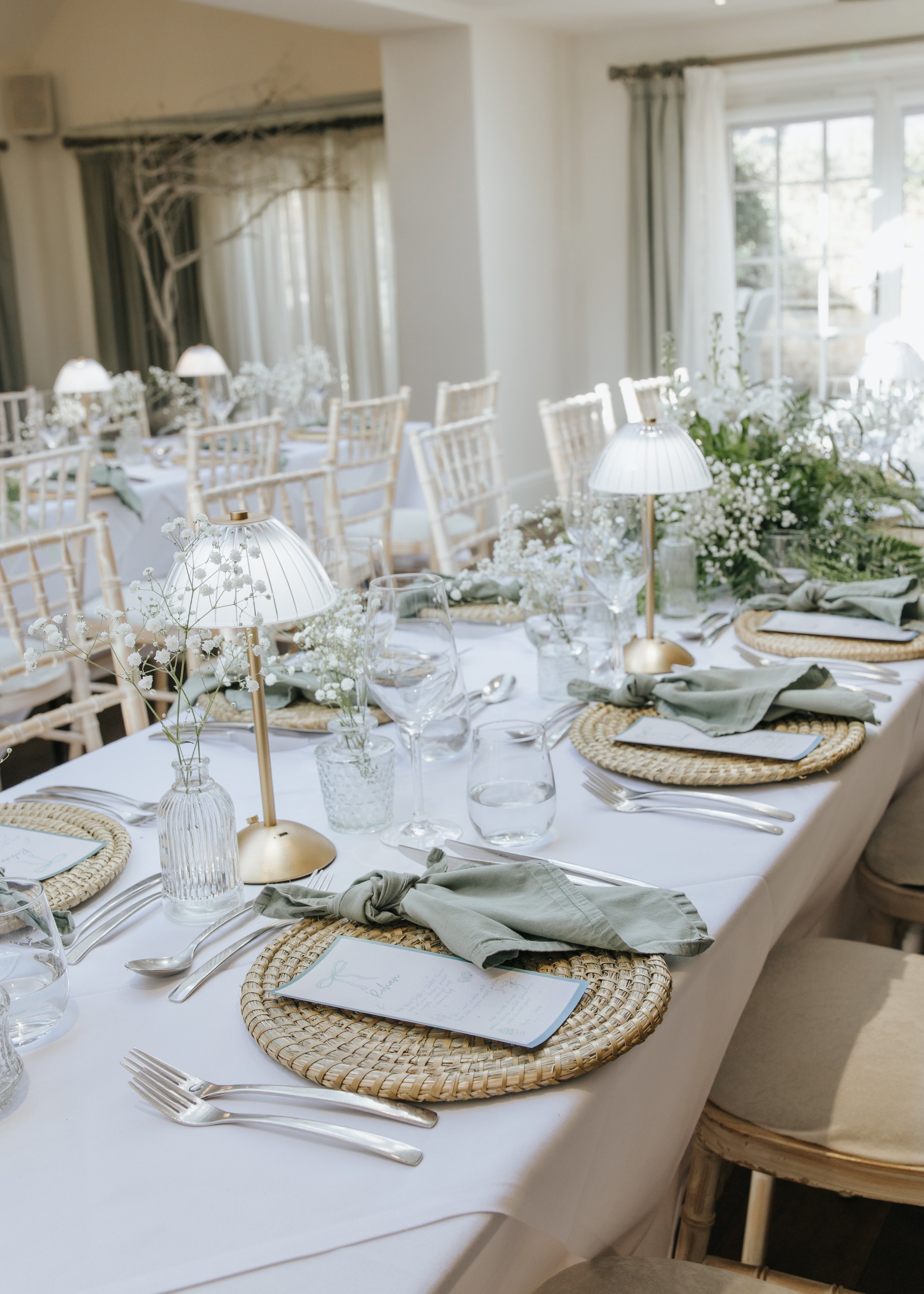 Elegant dining table decorated for a wedding or special event with white linens, wicker chargers, green napkins, floral centerpieces, and small table lamps, with chairs and windows in the background.