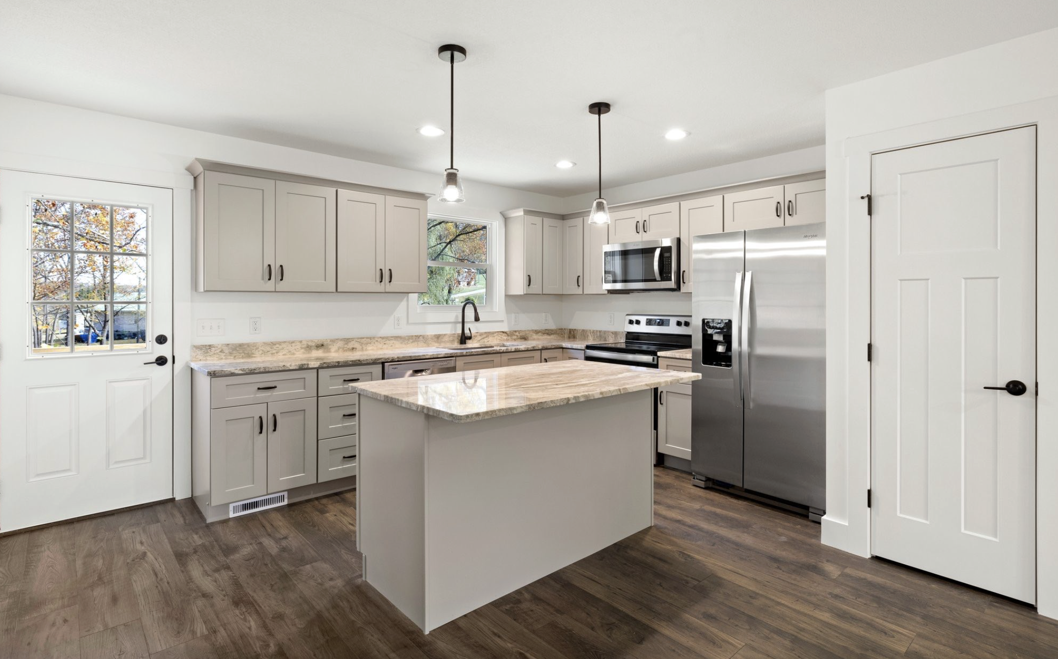Modern kitchen with gray cabinets, granite countertops, stainless steel appliances, and wooden flooring, with a door and window showing outdoor trees.