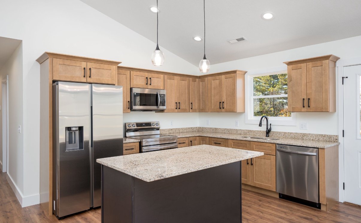 Modern kitchen with light wooden cabinets, stainless steel refrigerator, microwave, stove, dishwasher, granite countertops, a window above the sink, and two pendant lights.