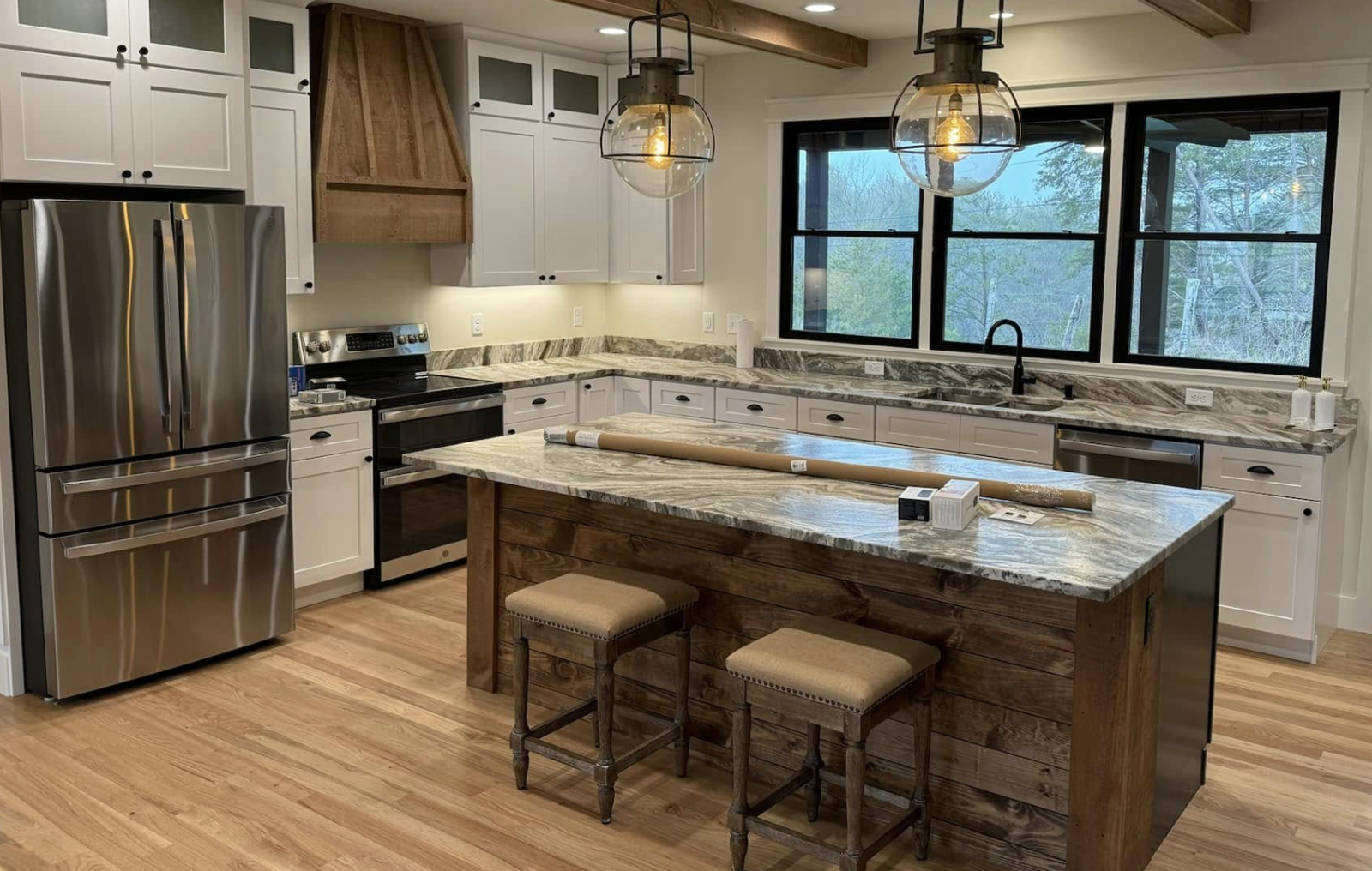 Modern kitchen with white cabinets, stainless steel refrigerator, black oven, marble countertops, and a wooden kitchen island with two stools. Large window with black frames over the sink, hanging glass pendant lights, and hardwood flooring.