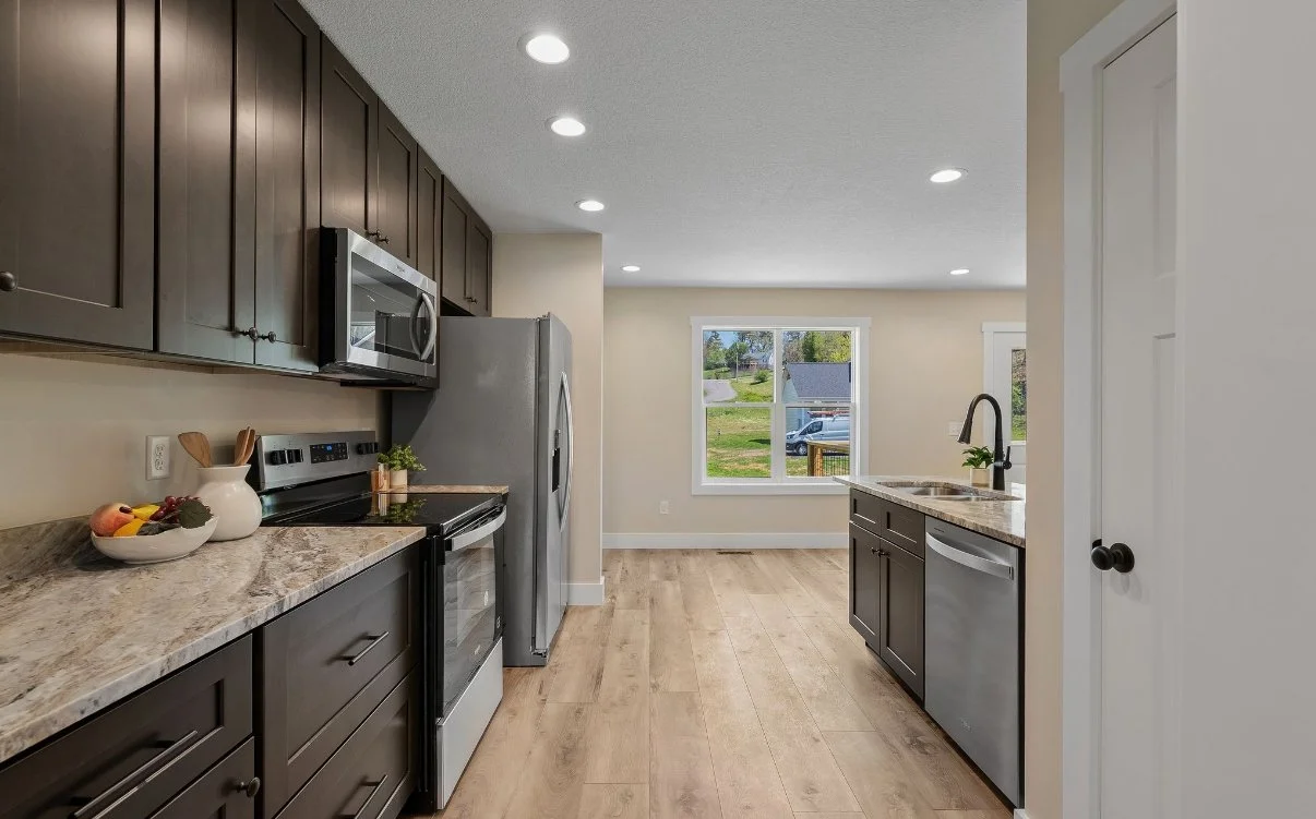 Kitchen with dark cabinets, stainless steel appliances, a center sink, light wooden flooring, and a large window overlooking a yard.