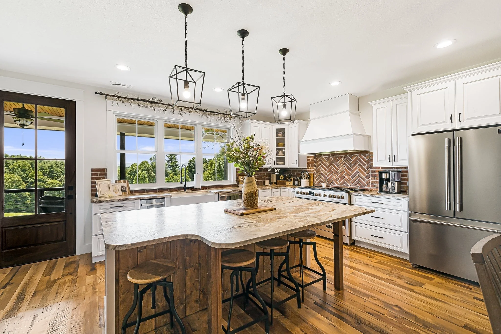 Modern kitchen with white cabinets, brick backsplash, large island with wooden top, stainless steel appliances, and a large window showing greenery outside.