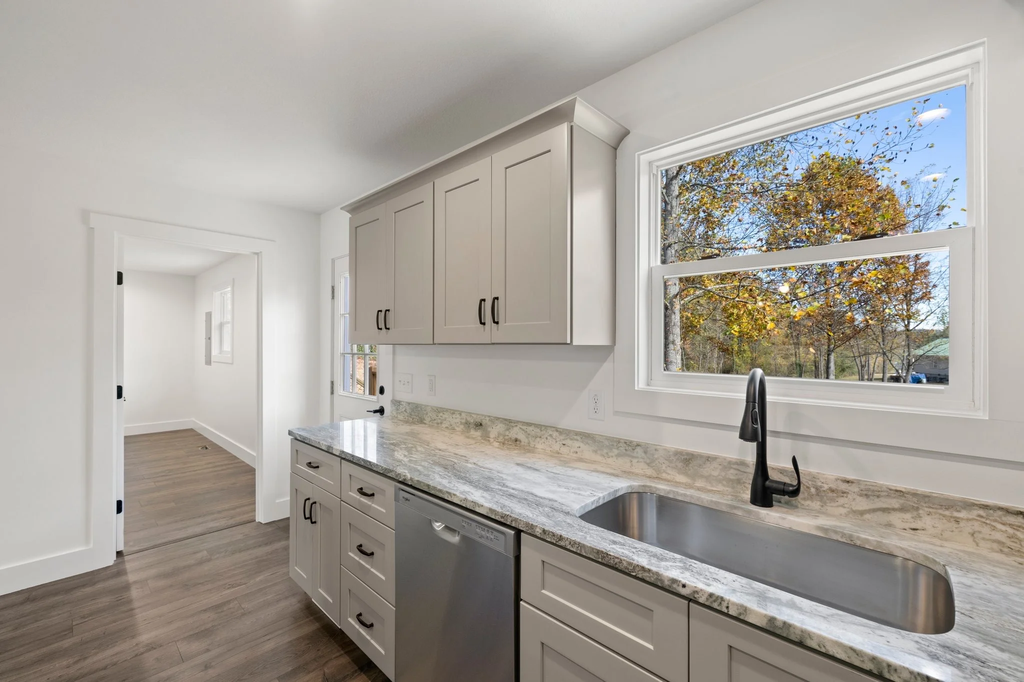 Kitchen with white cabinets, granite countertops, stainless steel dishwasher, black faucet, large window showing trees with autumn leaves, and a doorway leading to another room.