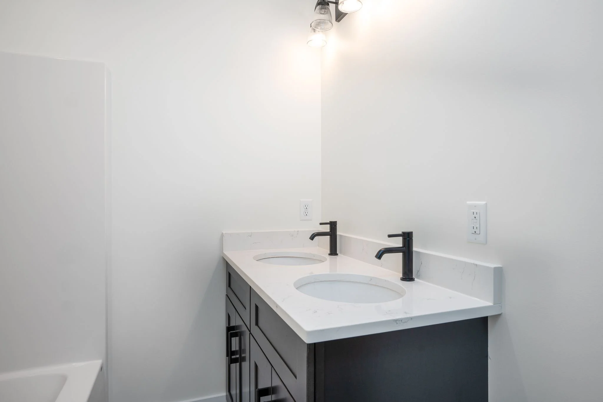 Modern bathroom vanity with double sinks, black faucets, and a white marble countertop, against a white wall with electrical outlets and a light fixture above.