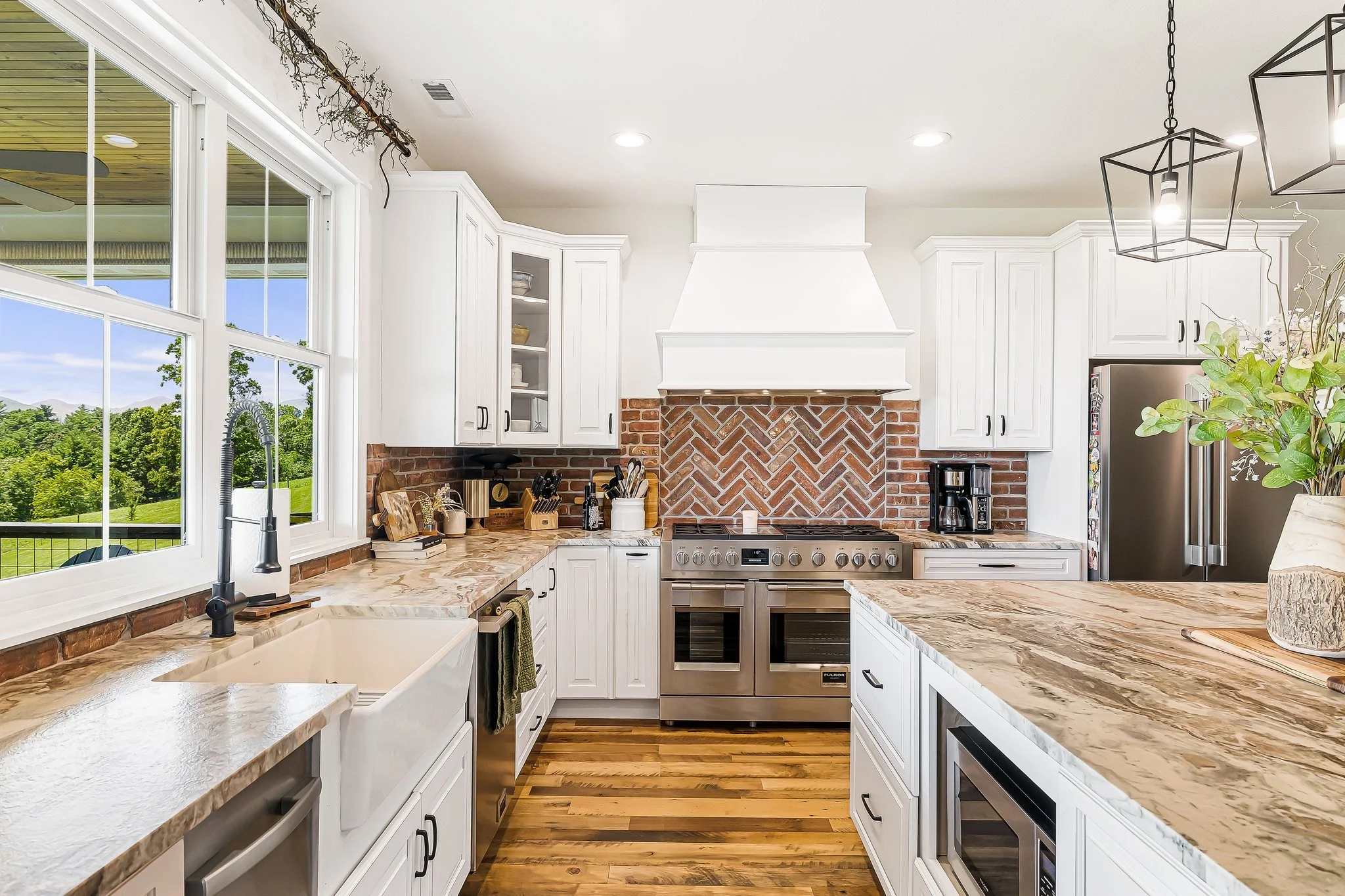 Modern kitchen with white cabinets, brick backsplash, wooden countertops, stainless steel appliances, large windows showing a green landscape, and decorative black pendant lights.