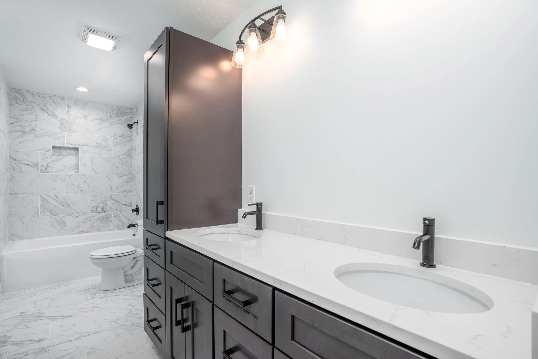 Modern bathroom with double vanity sinks, dark wood cabinets, white marble countertops, and a marble-tiled bathtub/shower area with black fixtures.