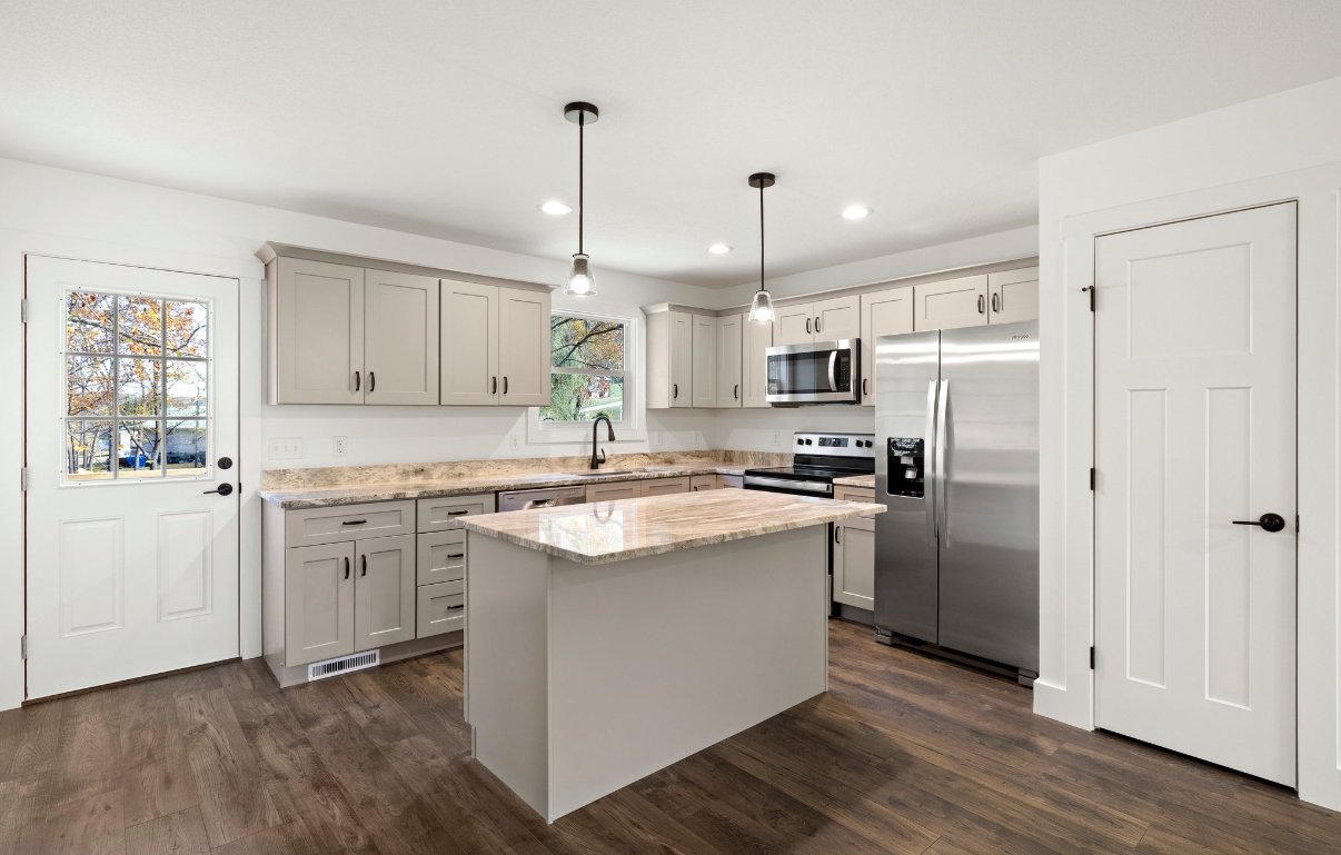 Modern kitchen with gray cabinets, stainless steel refrigerator, microwave, and oven, granite countertops, small island, hardwood floors, and two pendant lights hanging from the ceiling.