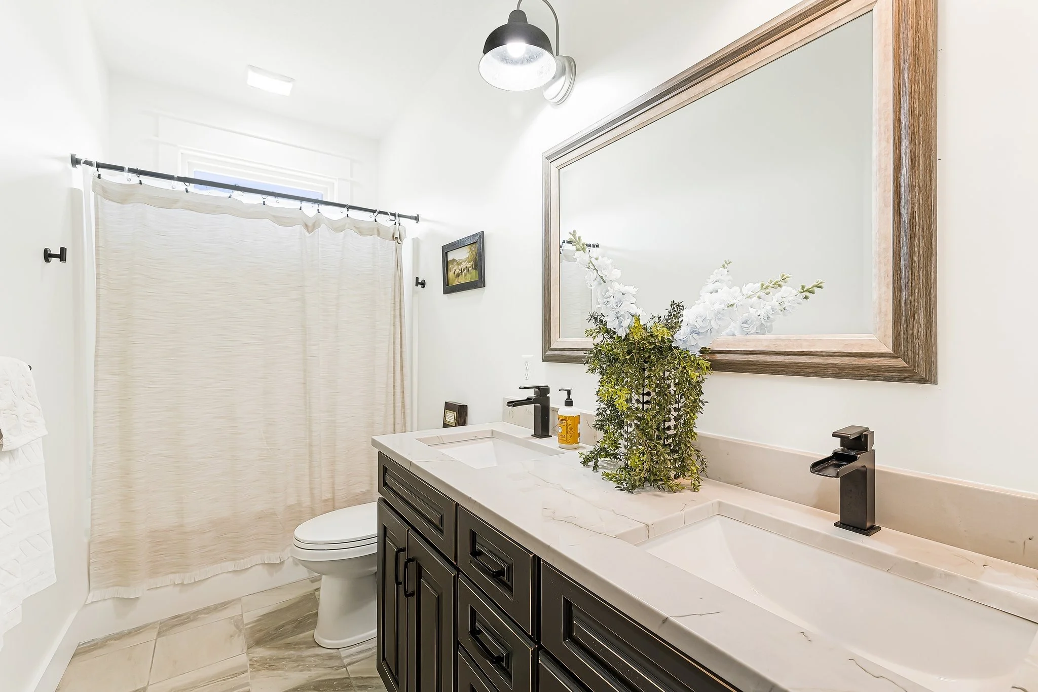 Modern bathroom with double vanity, large mirror, white countertop, and black fixtures, beige shower curtain, and marble floor tiles.