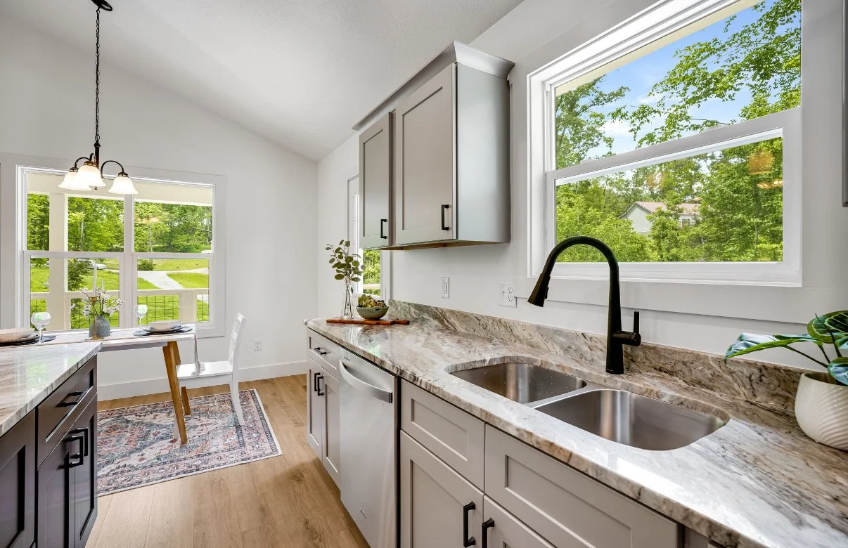 Kitchen with granite countertops, white cabinets, and a double sink. Large windows showing a green outdoor landscape. A hanging light fixture over the dining area and a small table with chairs.