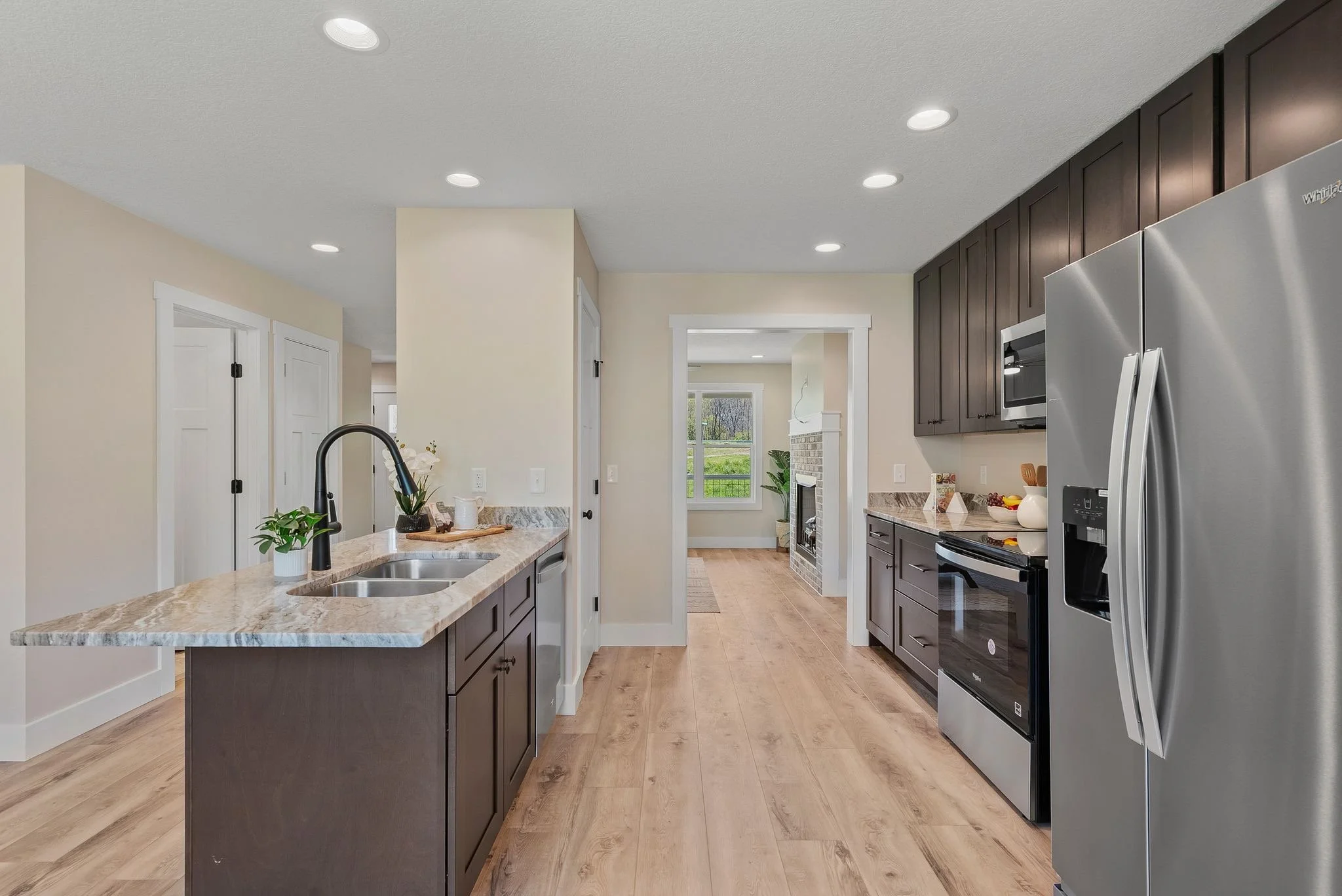 Modern kitchen with dark brown cabinets, stainless steel refrigerator, black microwave and oven, marble countertop island, light wood flooring, and a view into a living room with a fireplace and large window.