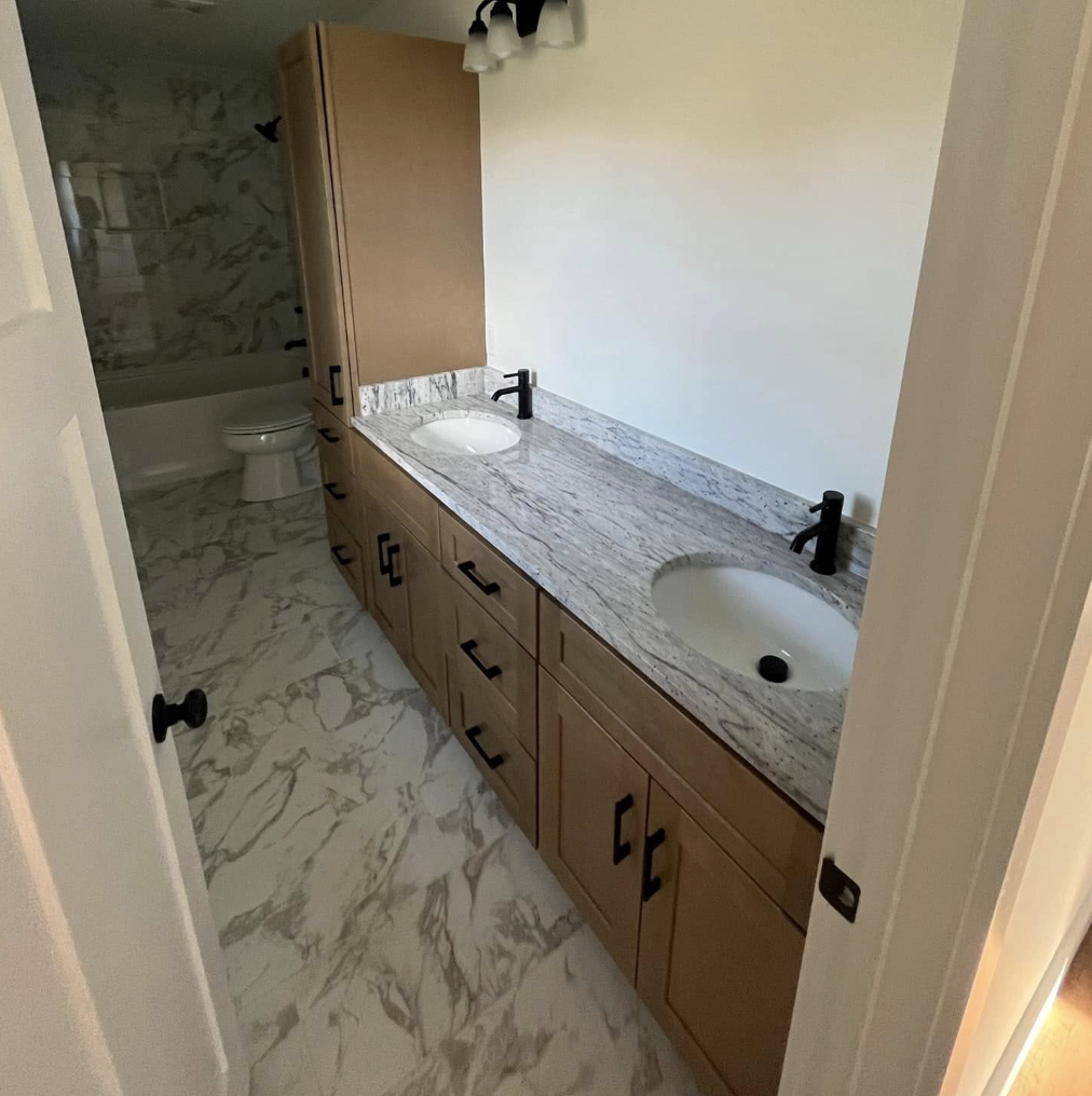 Bathroom with double vanity featuring a granite countertop, two undermount sinks with black faucets, and wooden cabinets with black handles. There is a marble floor and a partial view of a toilet and bathtub/shower area in the background.
