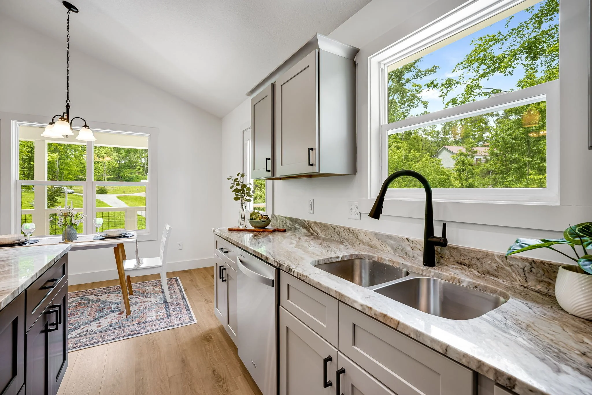 Bright kitchen with granite countertops, white cabinets, a double sink, and large windows revealing green trees outside.