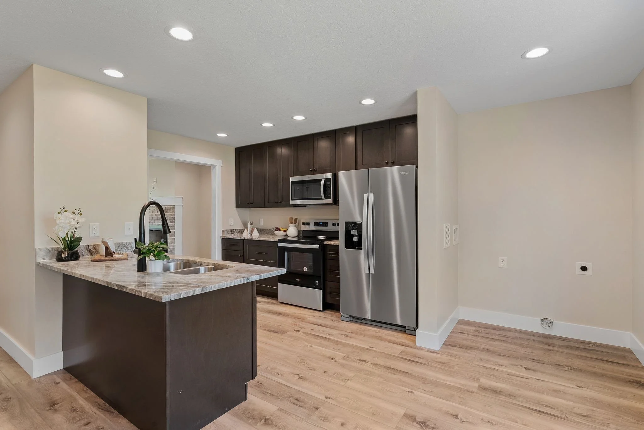 Modern kitchen with dark wood cabinets, stainless steel refrigerator, microwave, oven, and granite countertops. Light wood flooring and white walls. Small island with double sink, black faucet, and decorative plants.