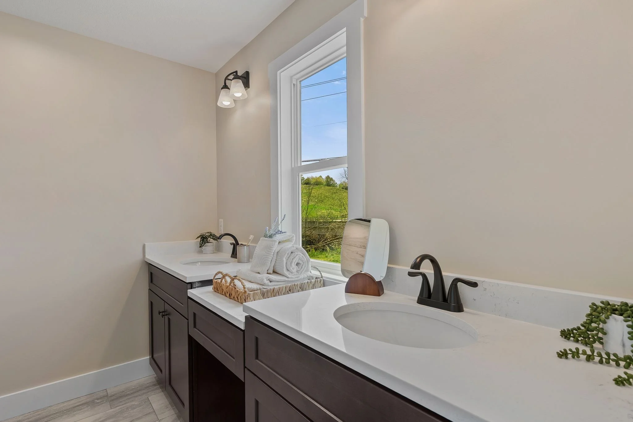 Bathroom double vanity with dark wood cabinets, white countertop, black faucet, towels, a small mirror, and a window showing green landscape and blue sky.