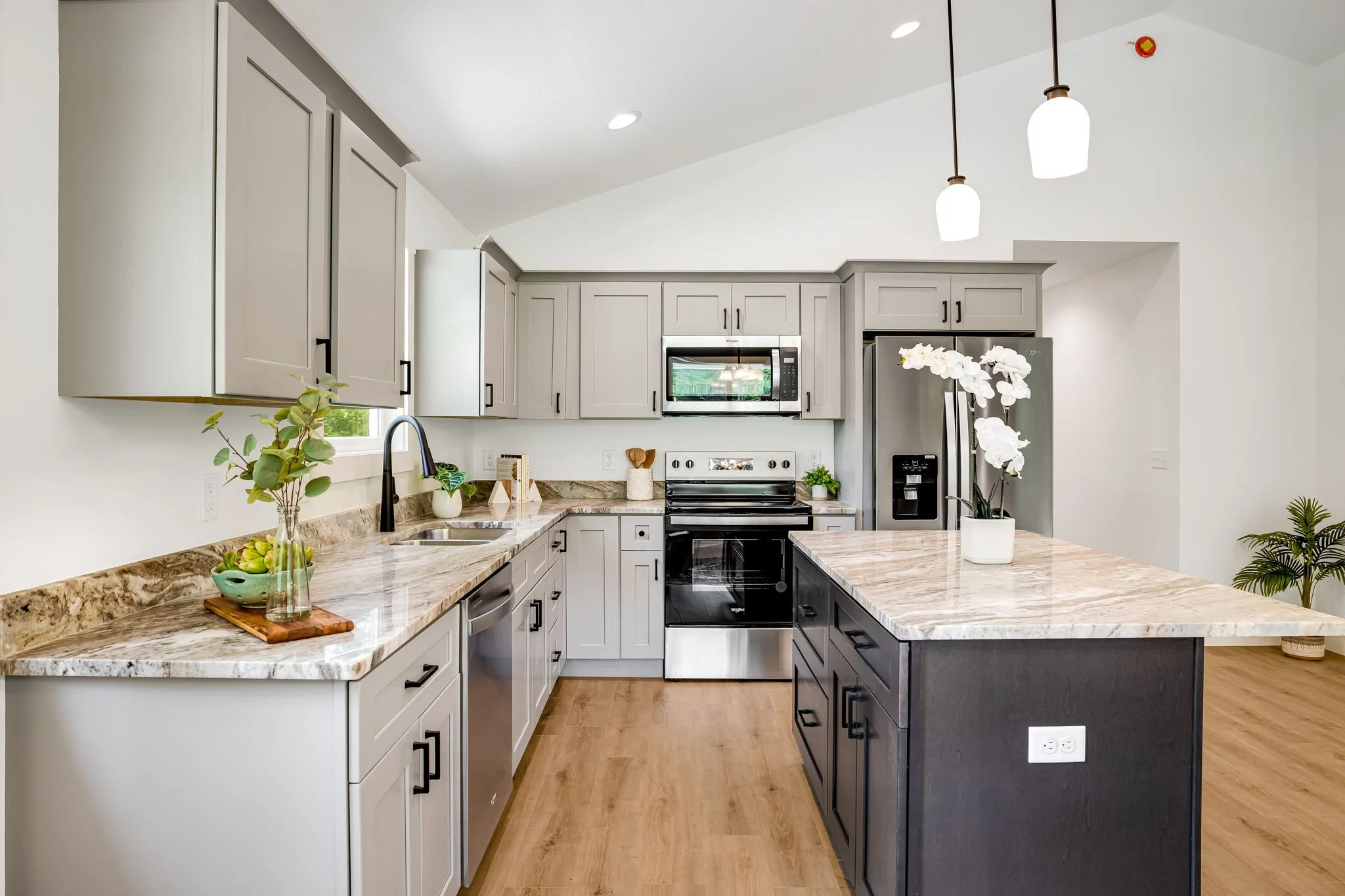 Modern kitchen with light gray upper cabinets, dark lower cabinets, marble countertops, stainless steel appliances, and a kitchen island with a marble top. There are pendant lights, a sink with a black faucet, and decorative plants and flowers.