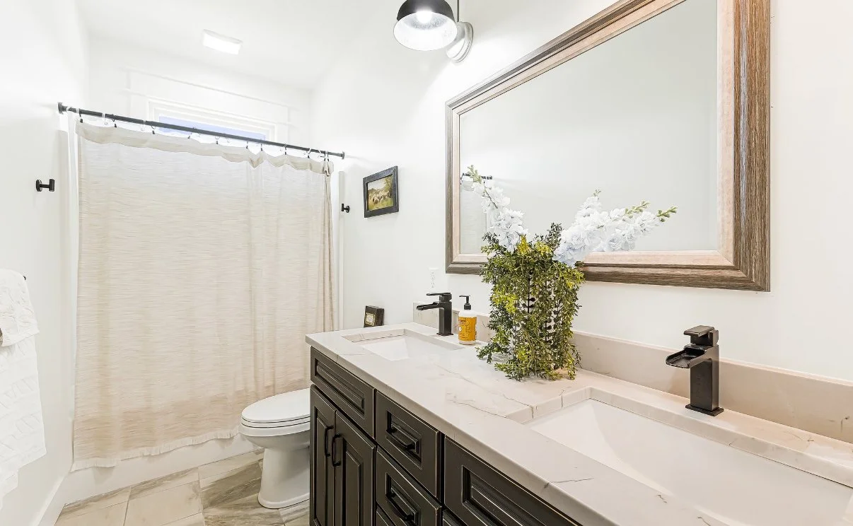 Bathroom with double sink vanity, large mirror, beige marble countertop, black fixtures, a potted plant, white shower curtain, small picture on the wall, and a toilet.