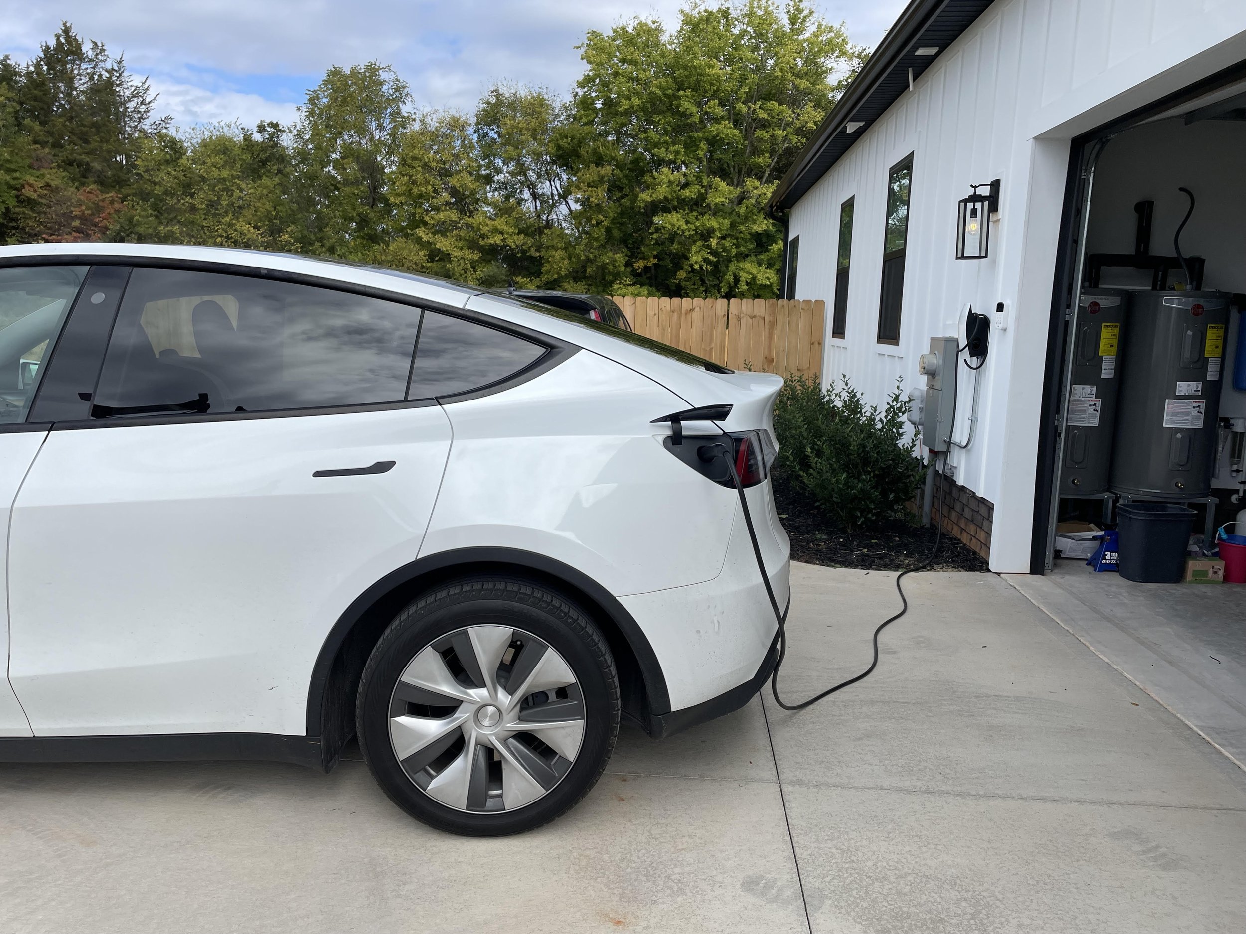 Side view of a white electric car plugged into a charger outside a white building with a garage door