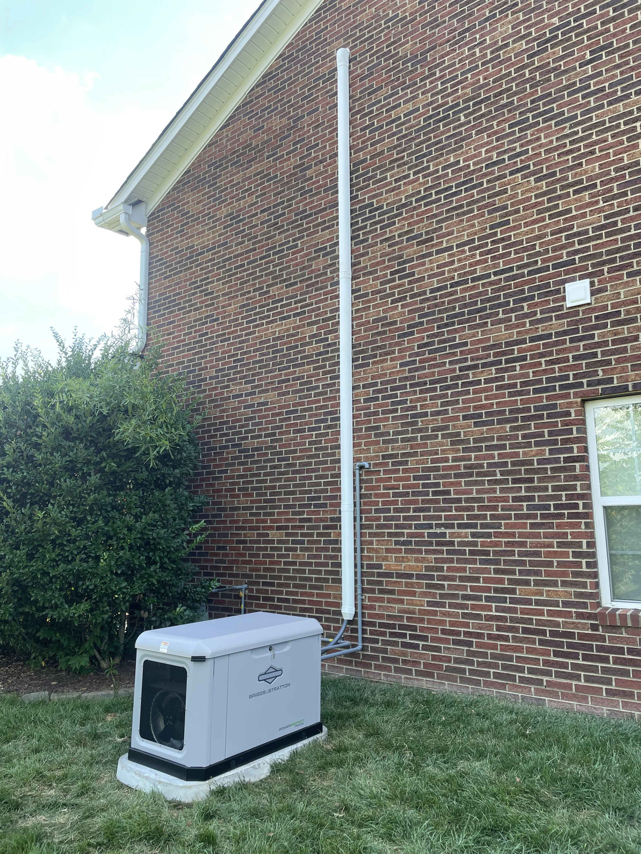 An outdoor view of a brick house wall with an underground electrical generator and a vertical conduit pipe running along the wall.