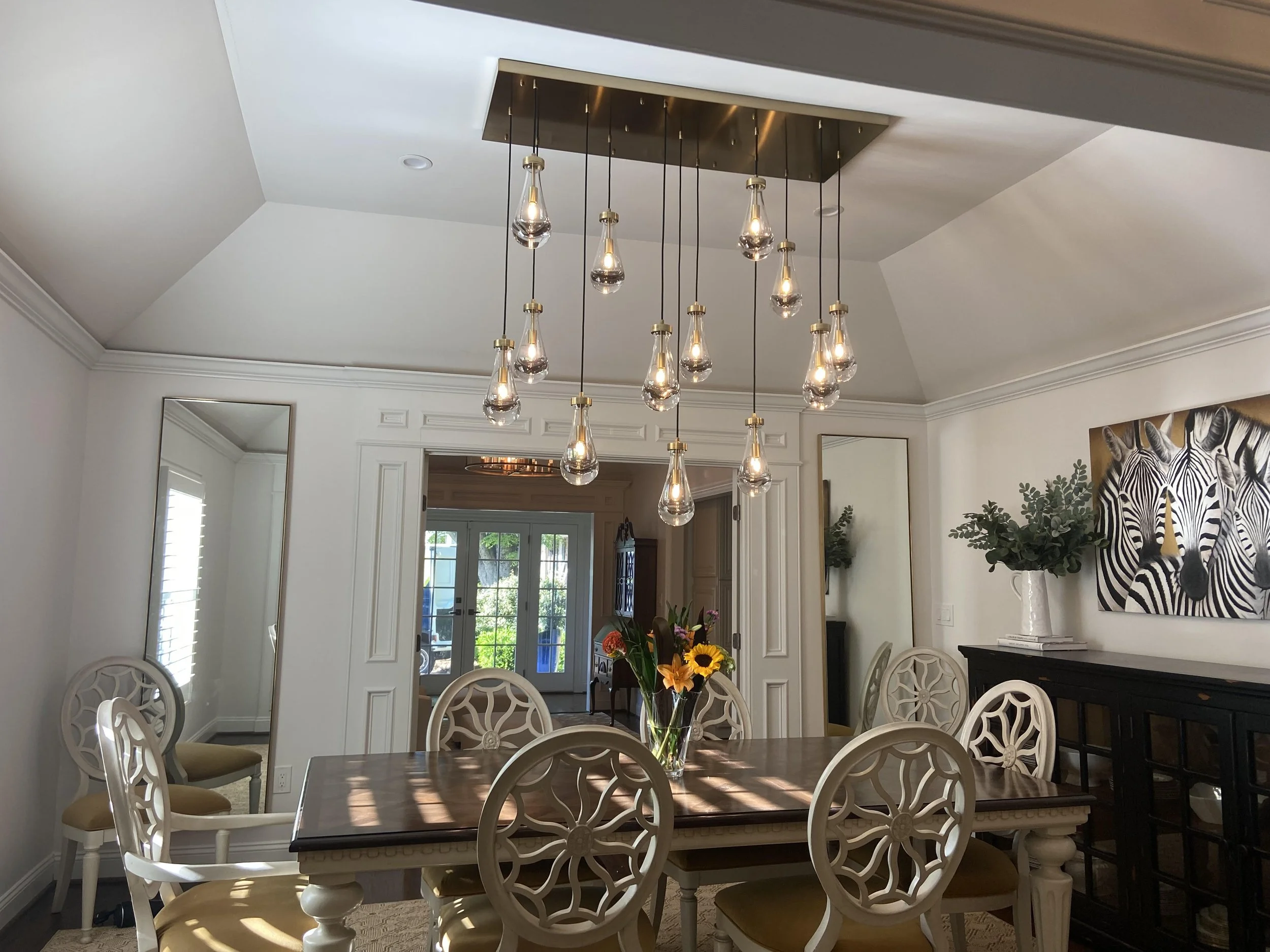 A dining room with a wooden table and cream-colored chairs with decorative backs. A vase of flowers is on the table. There are two large mirrors and a black sideboard with a vase of greenery. A zebra painting hangs on the wall. A ceiling light fixtur