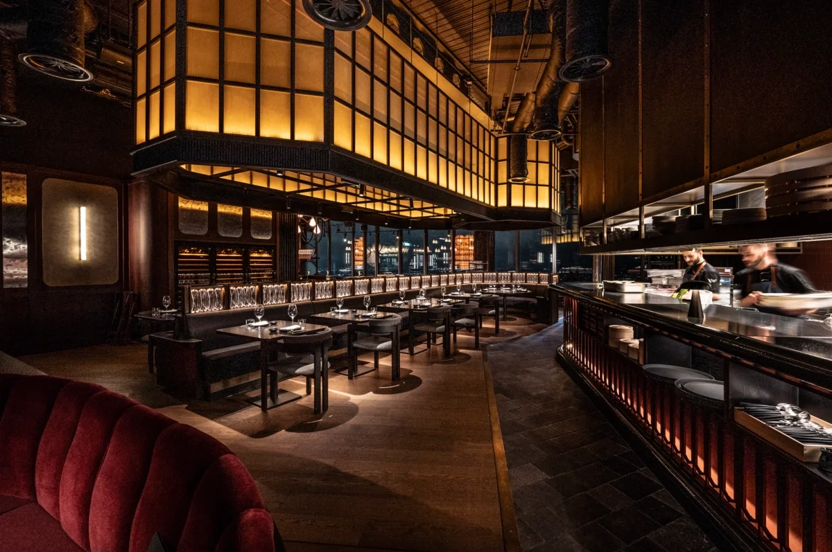 Elegant restaurant interior with dim lighting, dark wood accents, a long curved bench with glassware, and an open kitchen with chefs preparing food.