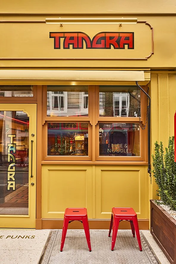 The storefront of a restaurant named TANGRA painted in yellow with a sign in red and black above the door. There are two red stools outside and a window with signs advertising snacks, chai, rice, and noodles.