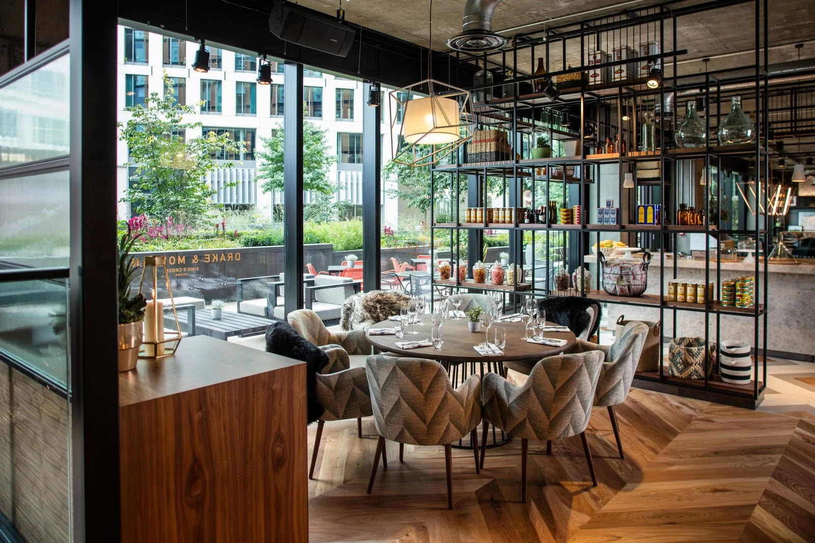 A modern restaurant interior featuring a round table with ten chairs, large glass windows showing an outdoor seating area, and decorative shelves with bottles and jars.