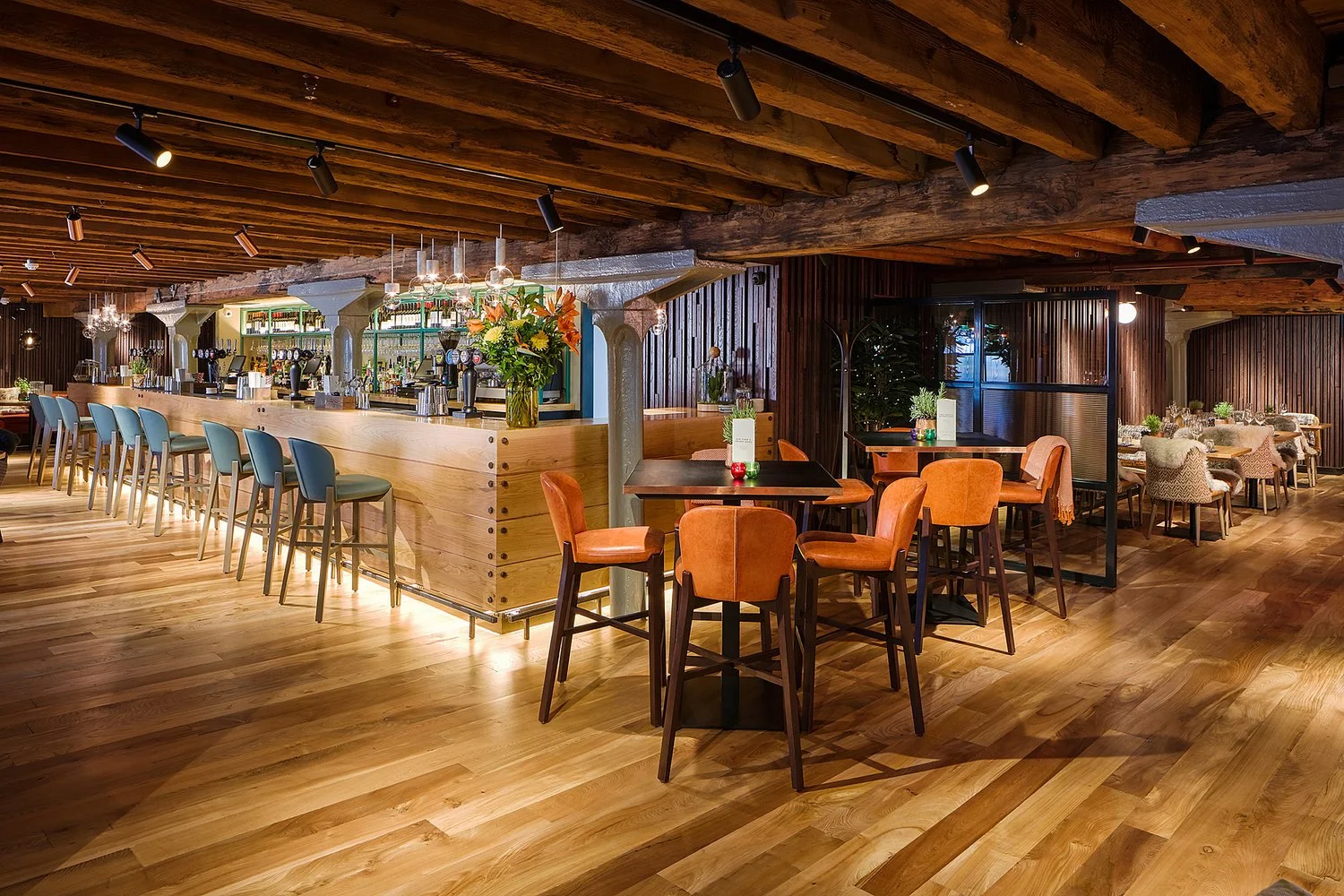 Interior of a cozy restaurant with wooden floors and ceiling beams, featuring a bar with blue chairs, and tables with orange chairs and decorative menus
