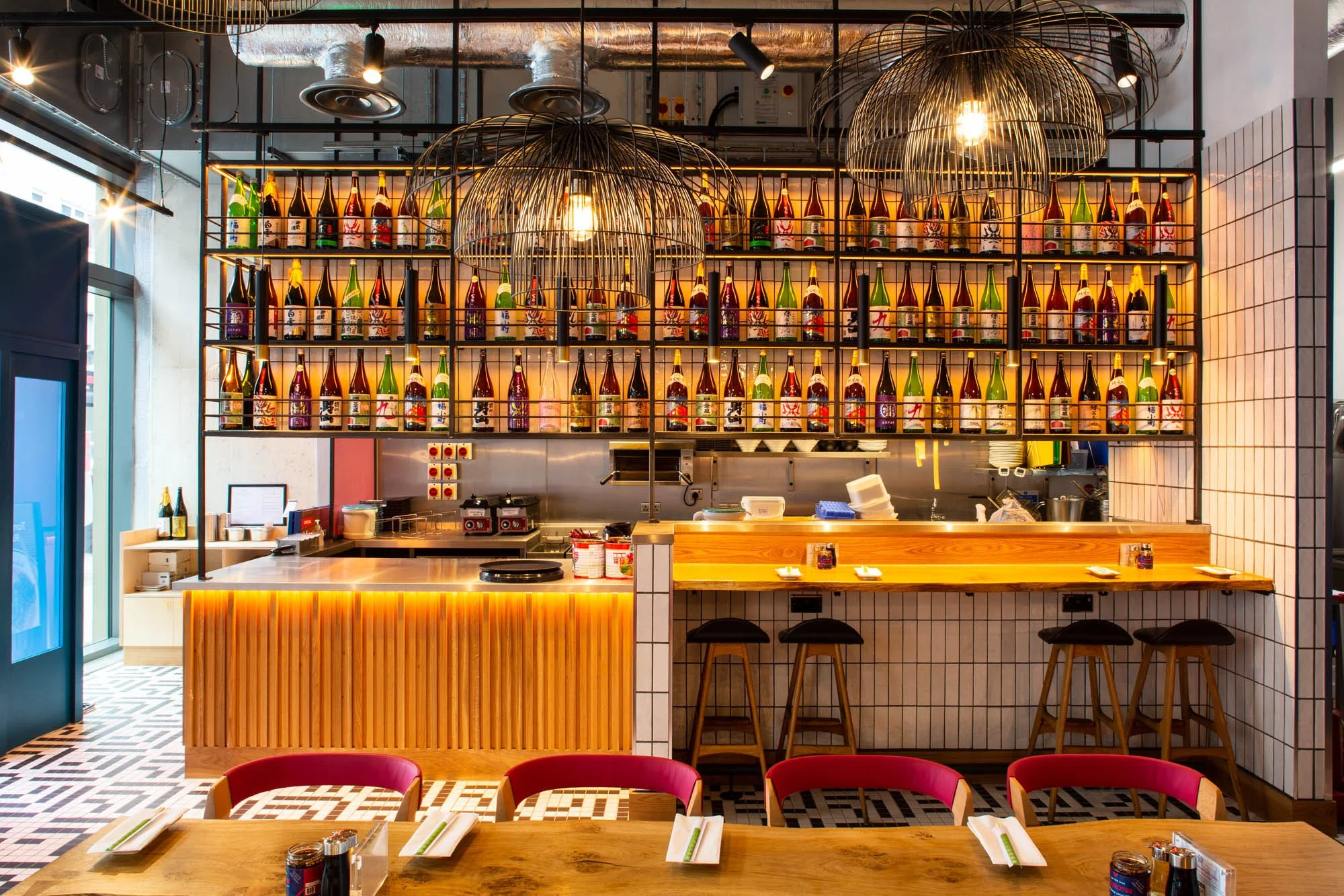 Interior of a modern Japanese restaurant or bar with a bar counter, bar stools, and shelves filled with colorful sake bottles. The decor includes wooden and tiled surfaces, black pendant lights, and an open kitchen area.